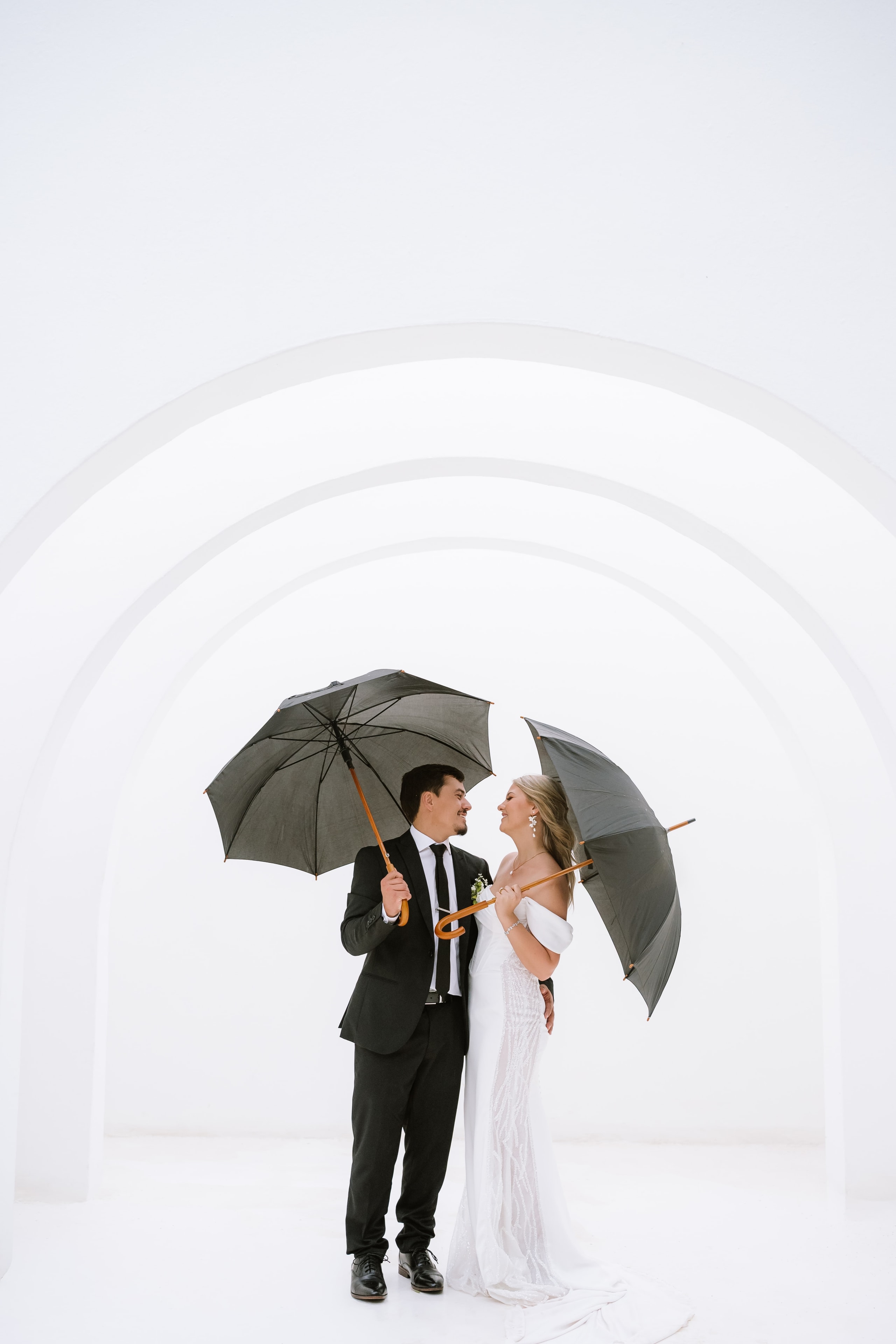 A bride and groom stand under umbrellas inside a bright white arched architectural structure