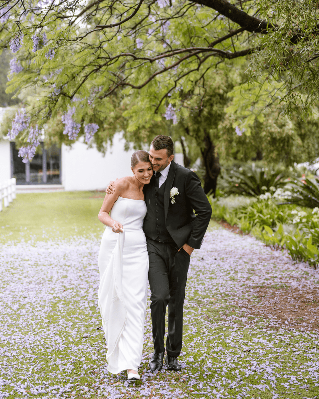 A newlywed couple walks hand in hand beneath blooming purple jacaranda trees in South Africa