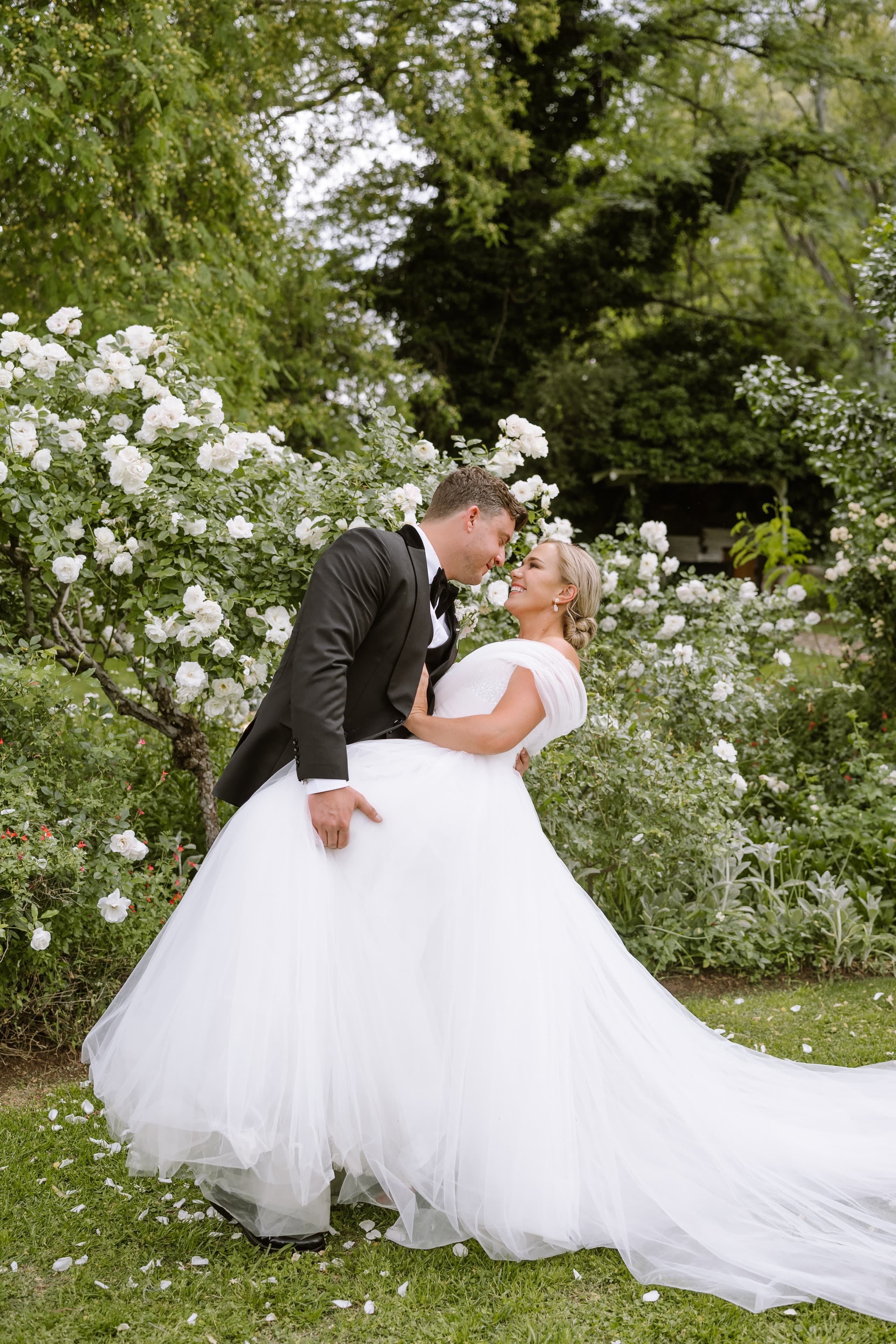 A groom in a black tuxedo lifts his bride in a white gown outdoors