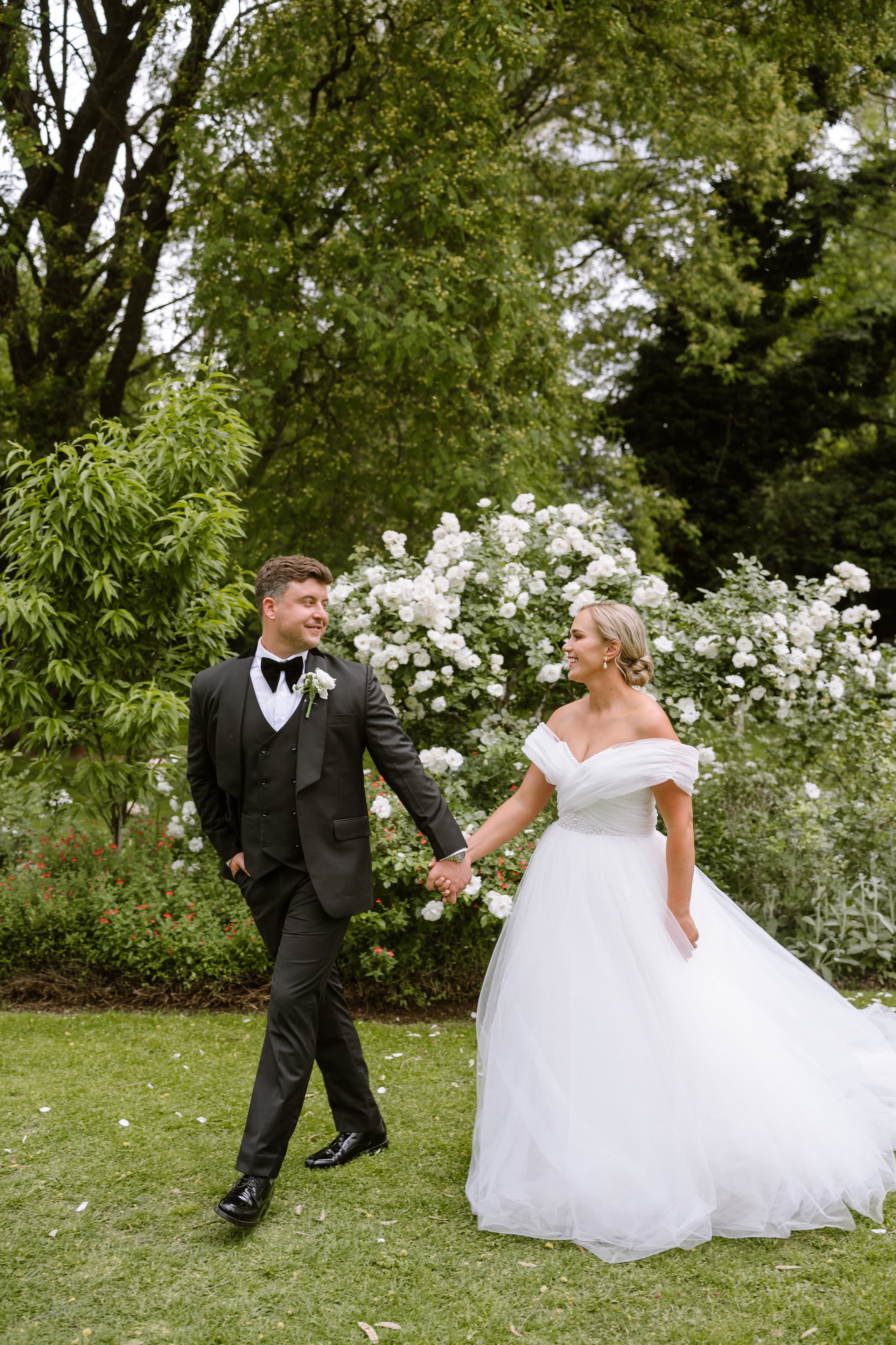 A newlywed couple walks hand in hand through a lush garden filled with white flowers