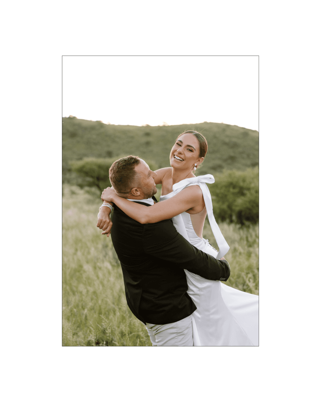 A groom lifts his smiling bride in a romantic embrace within a grassy field landscape