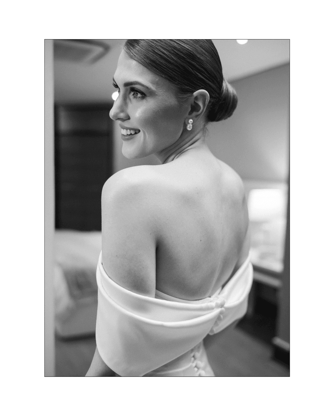 A smiling bride in an off-the-shoulder wedding dress looking over her shoulder in black and white