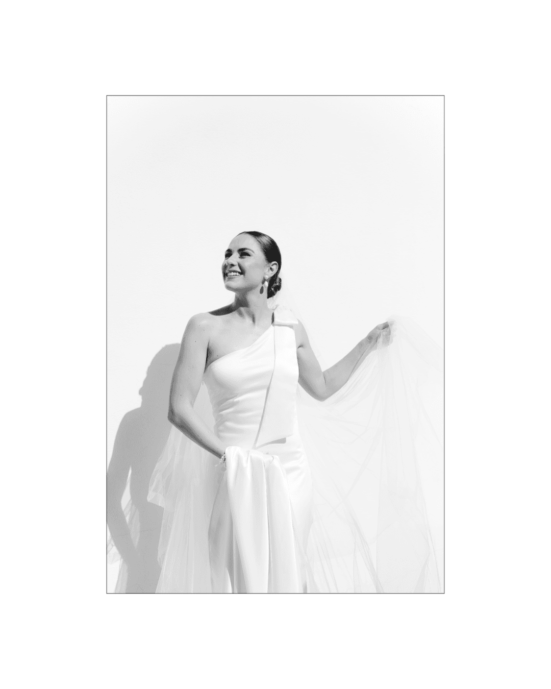 A smiling bride in a white one-shoulder gown holds her veil against a bright wall