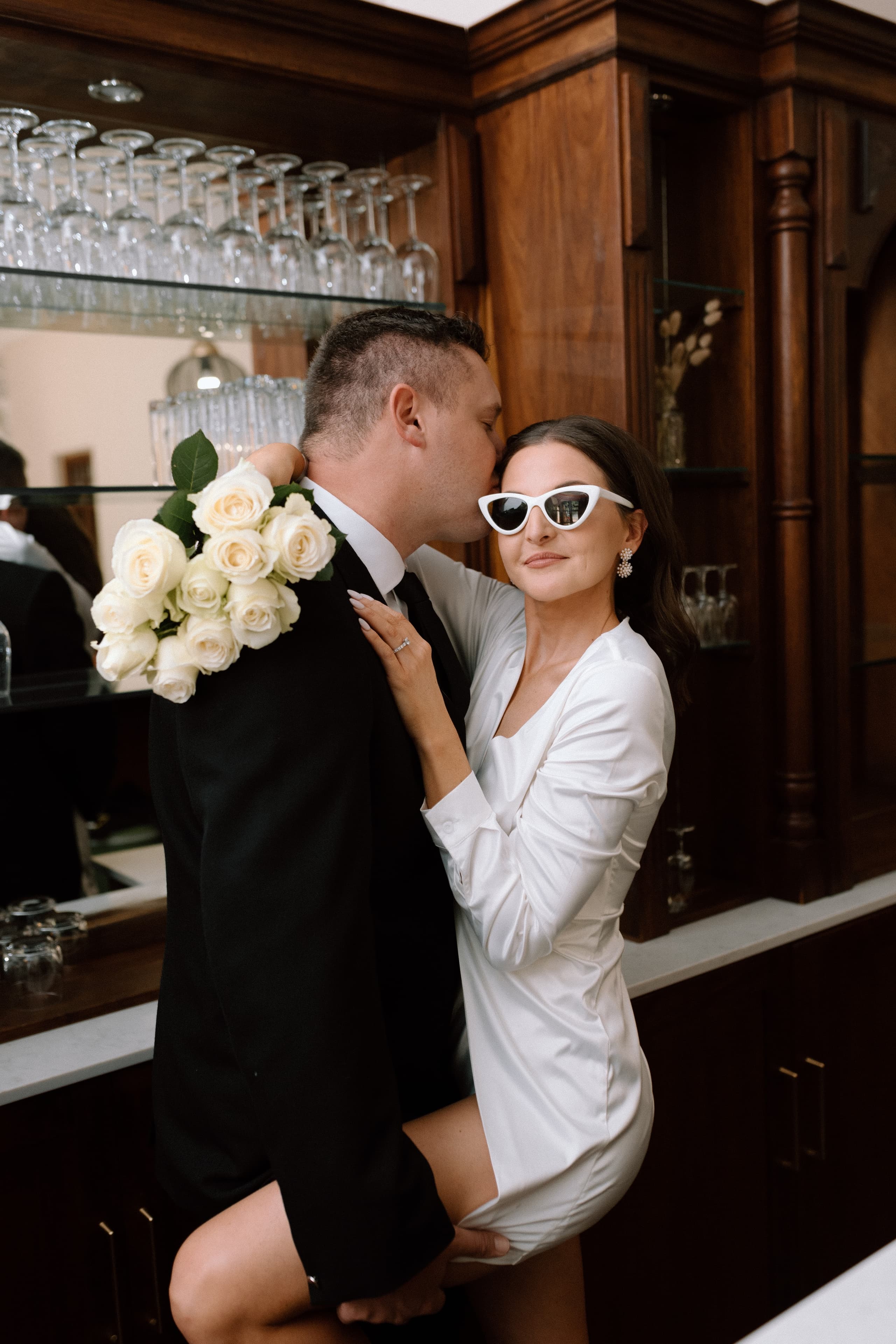 A stylish bride wearing white sunglasses rests her leg on her groom in a bar