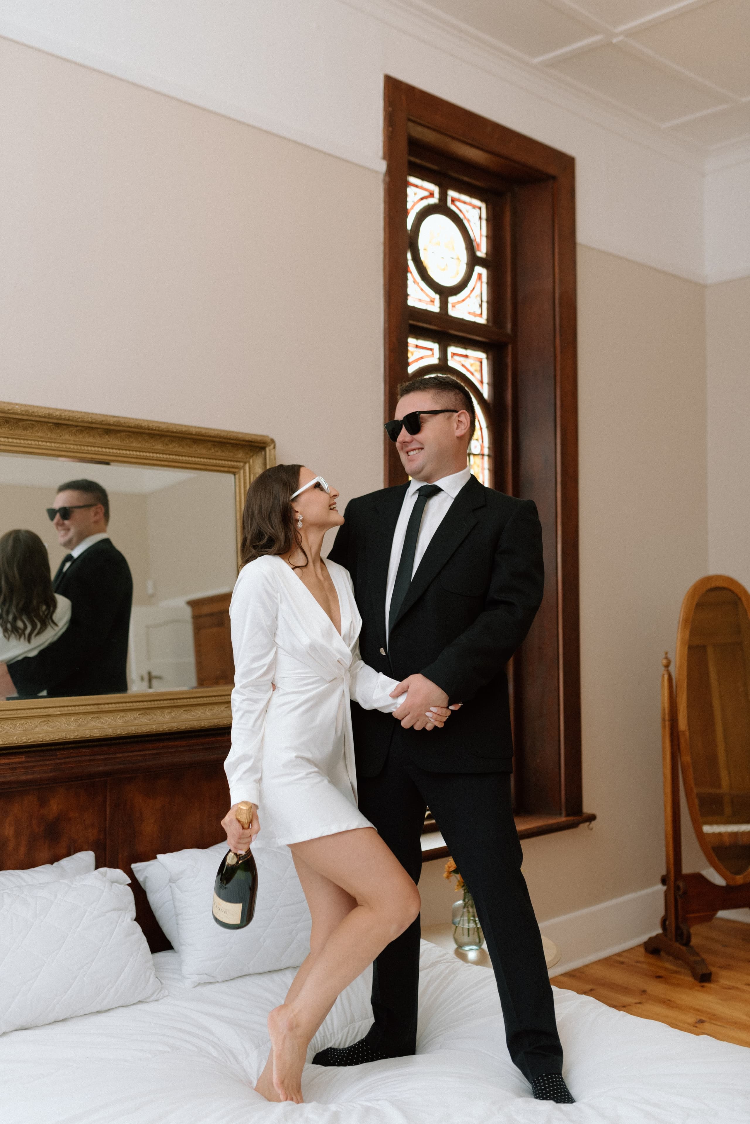 A stylish couple in sunglasses celebrates on a bed with a bottle of champagne