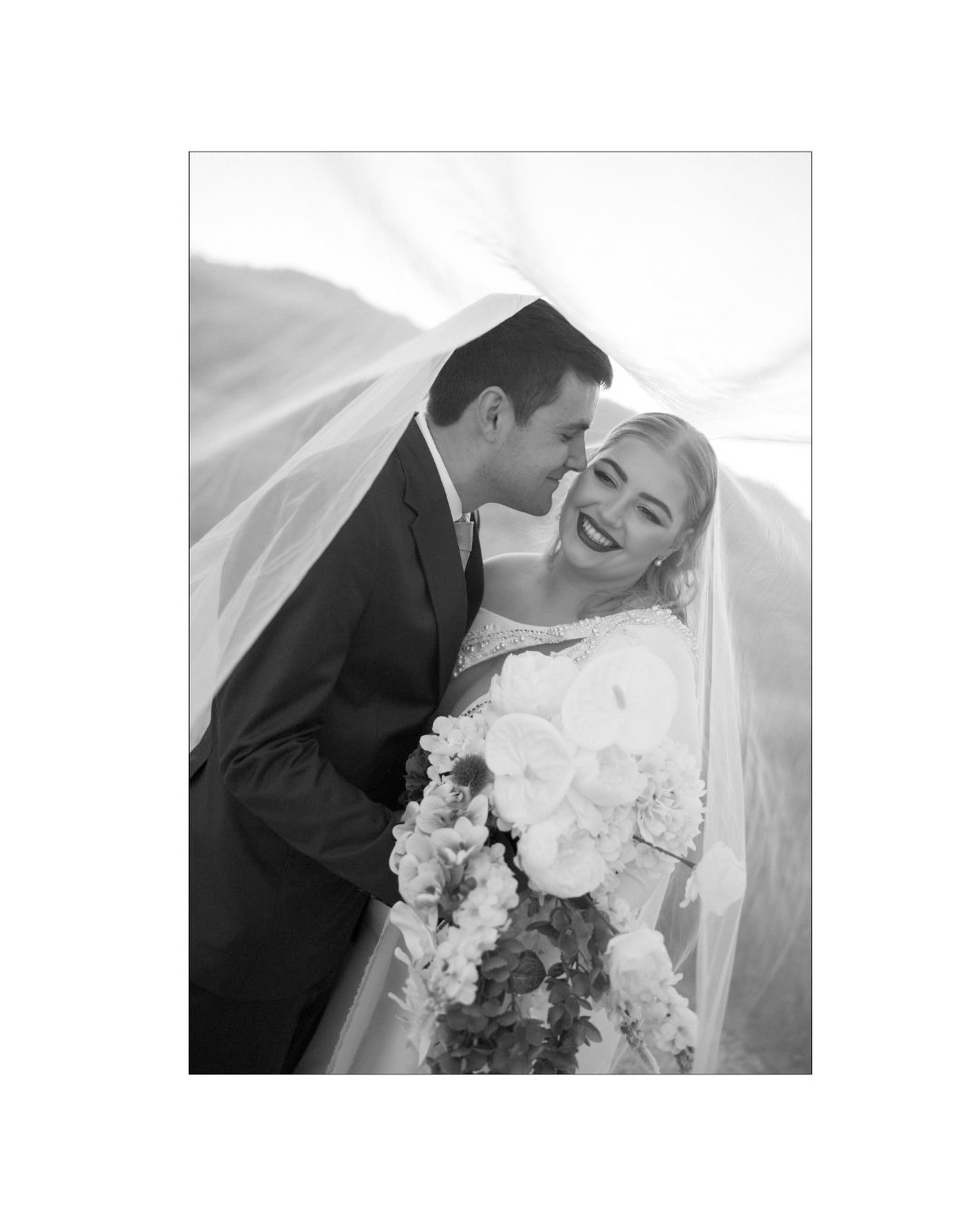 A black and white portrait of a bride and groom under a flowing veil