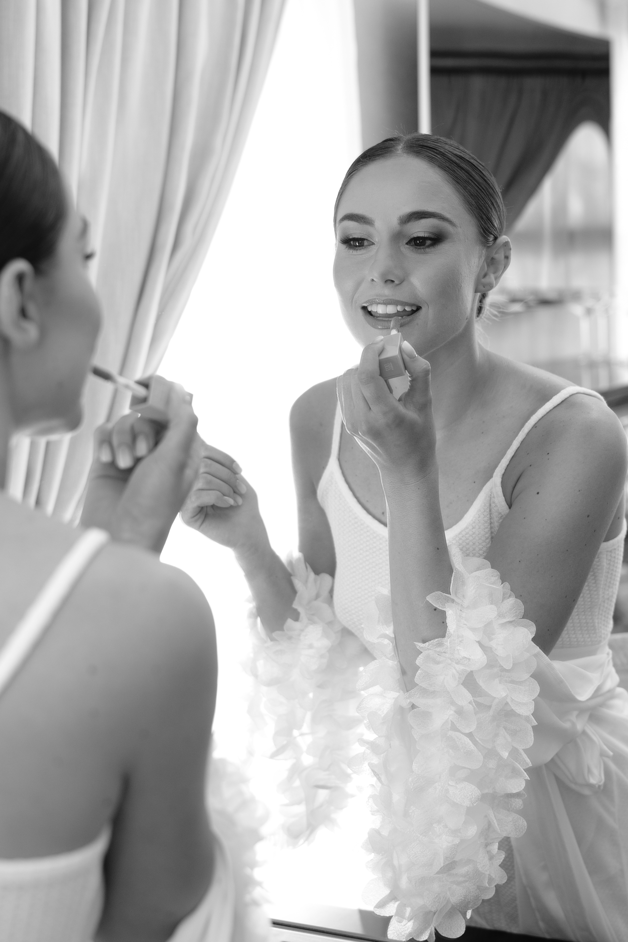 A bride in a white robe applies lipstick while looking at her reflection in a mirror