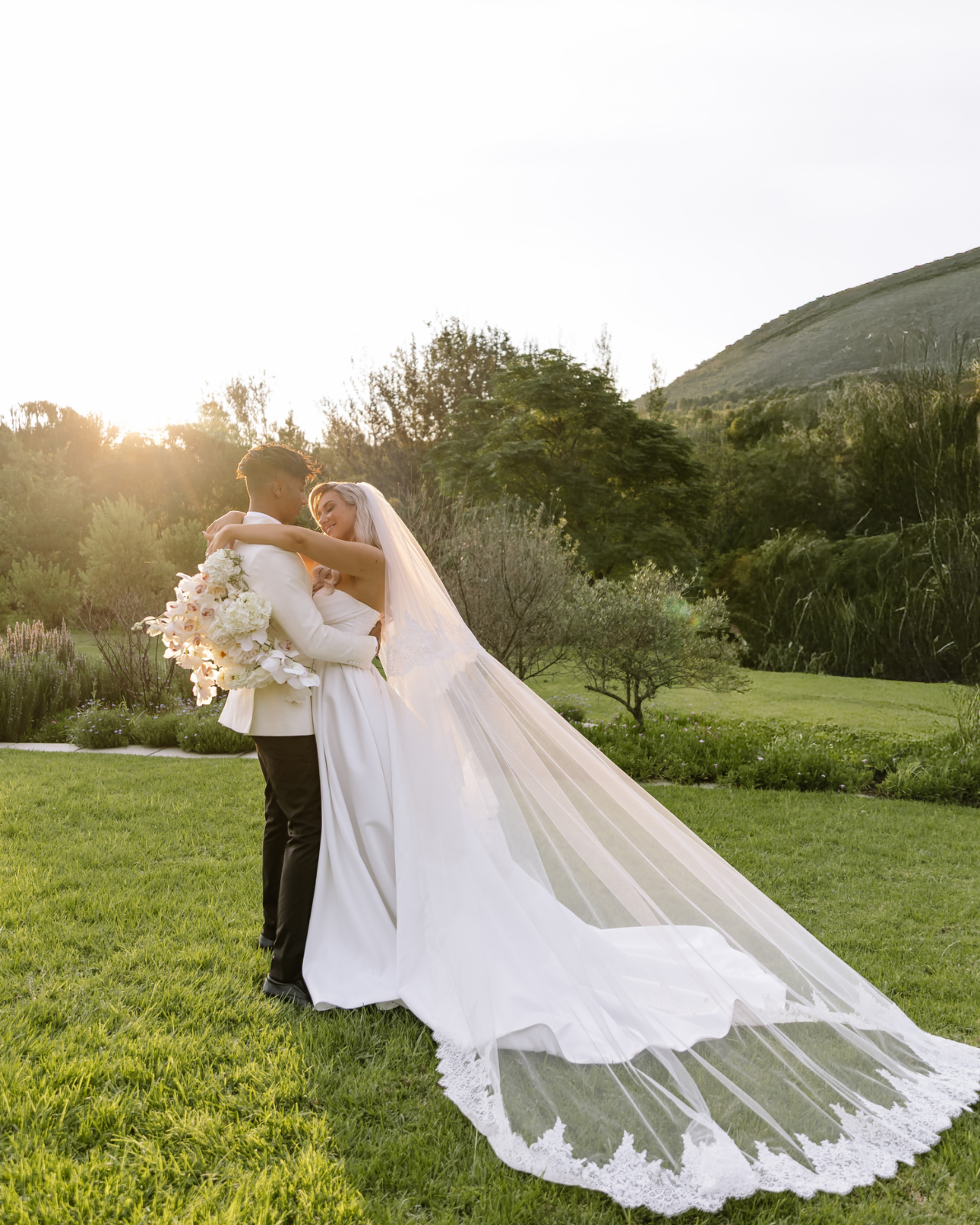 A bride and groom embrace in a sunlit garden during their luxury wedding portrait session