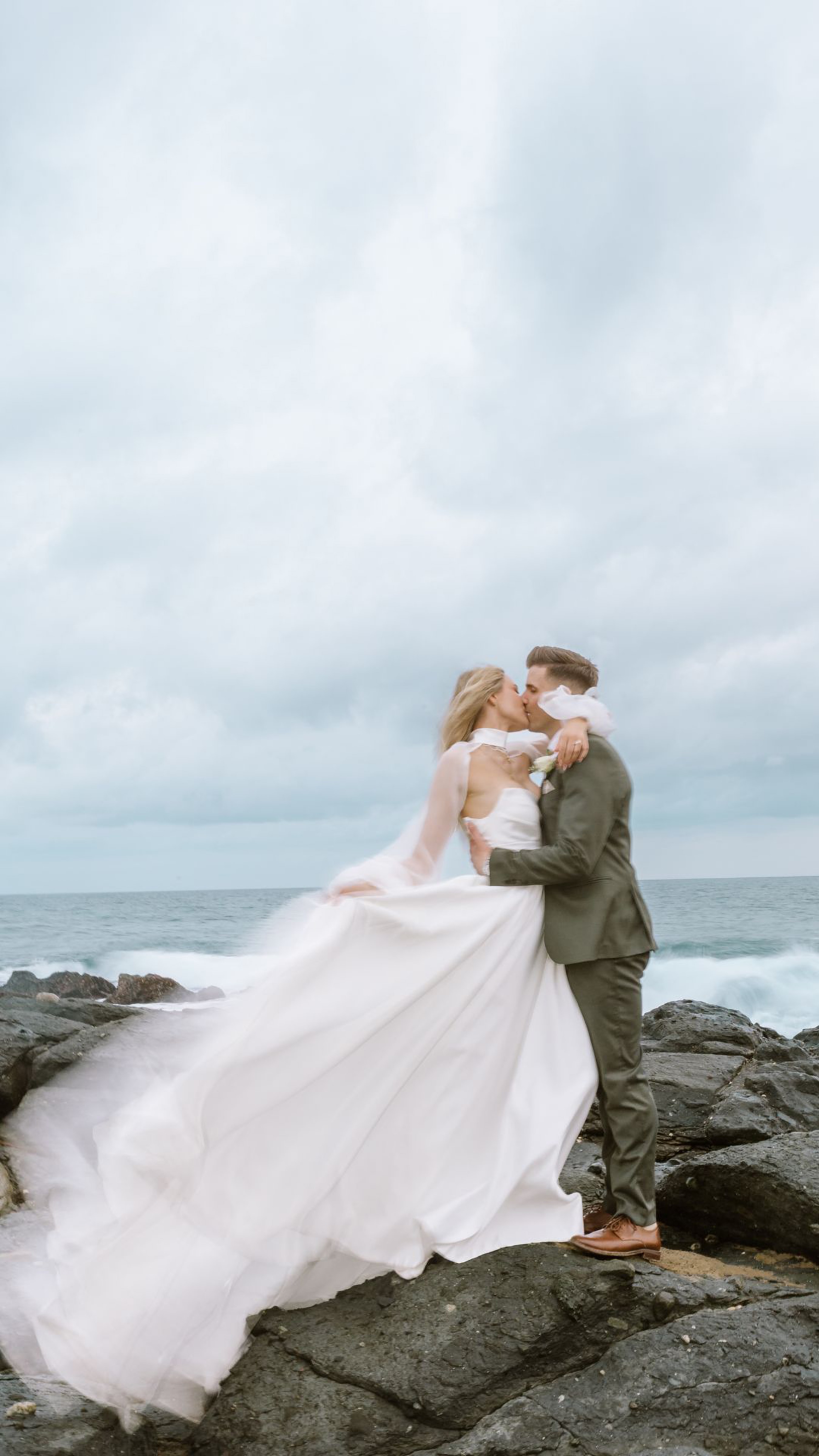 A bride and groom kiss on rocky ocean shore under a dramatic cloudy sky