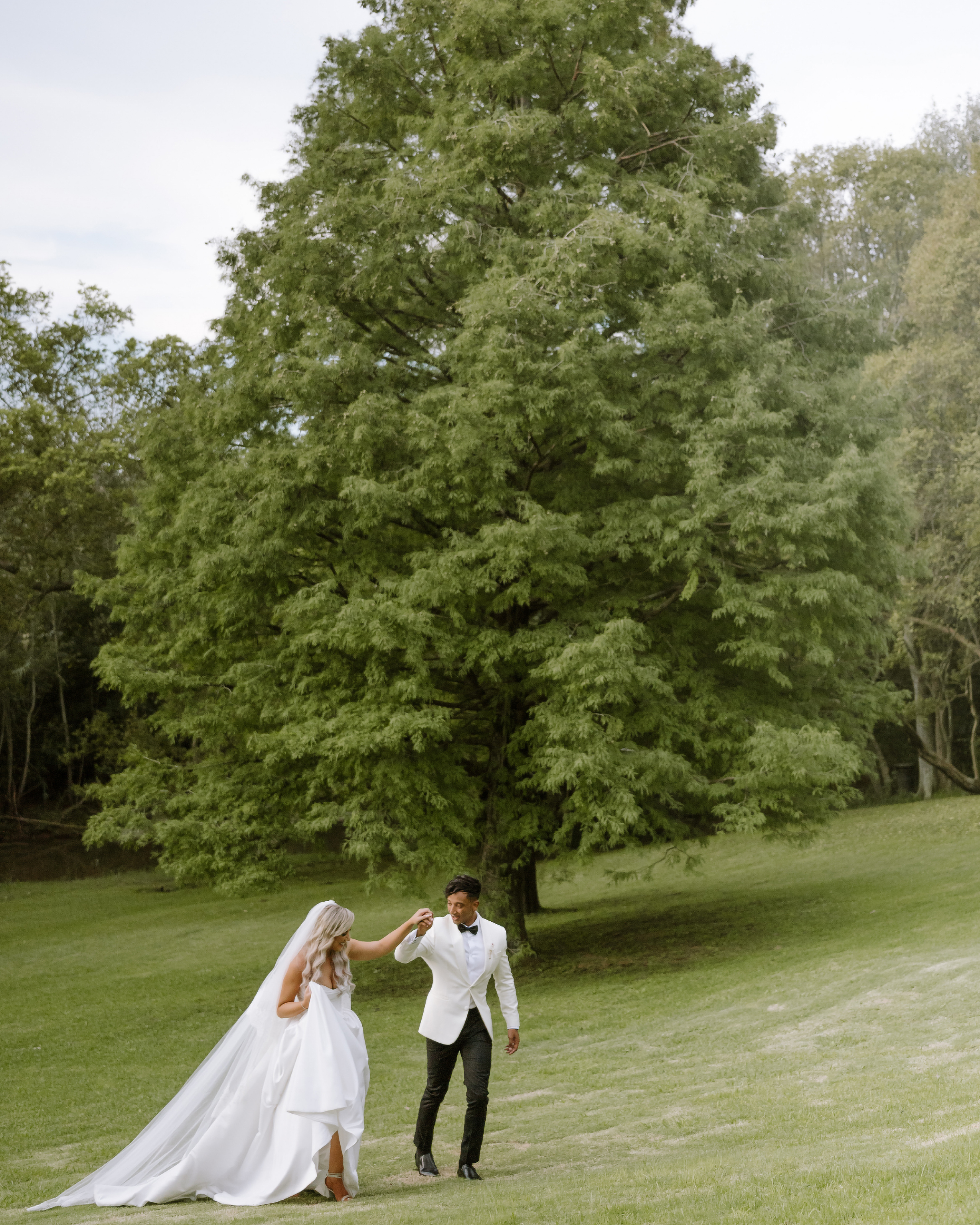 A bride and groom walk hand in hand across a lush green meadow outdoors