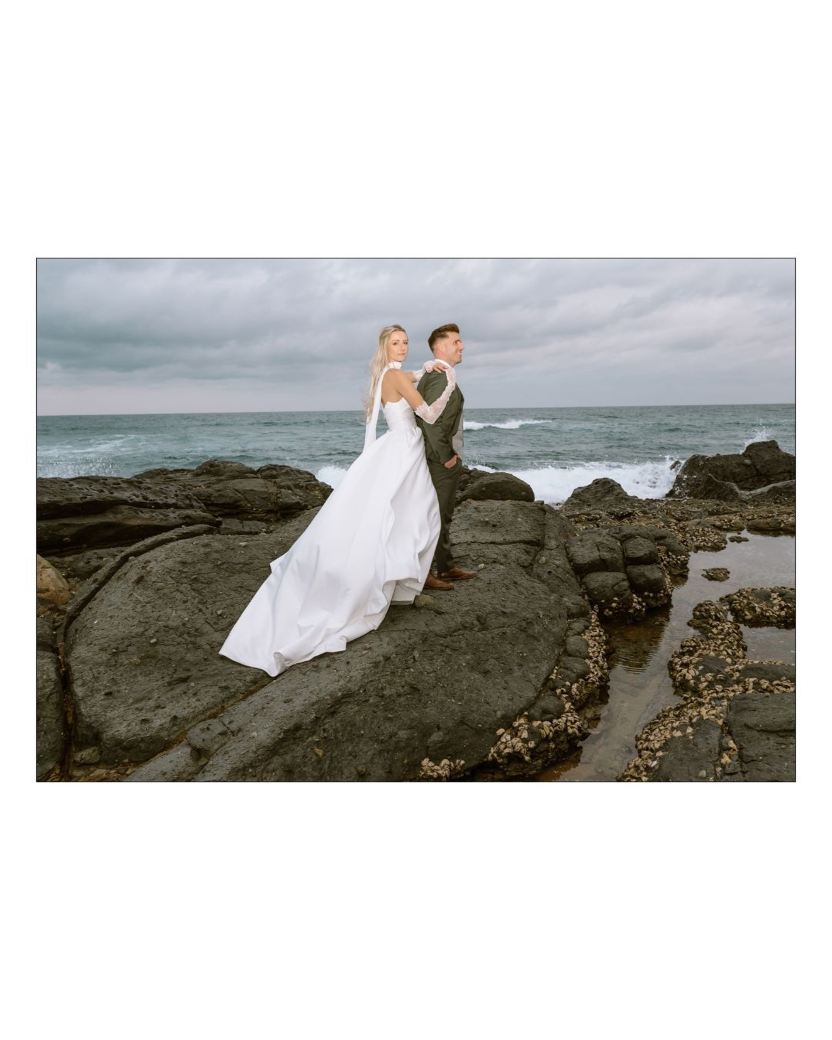 A bride in a white gown and groom standing on rocky coastal shores looking seaward