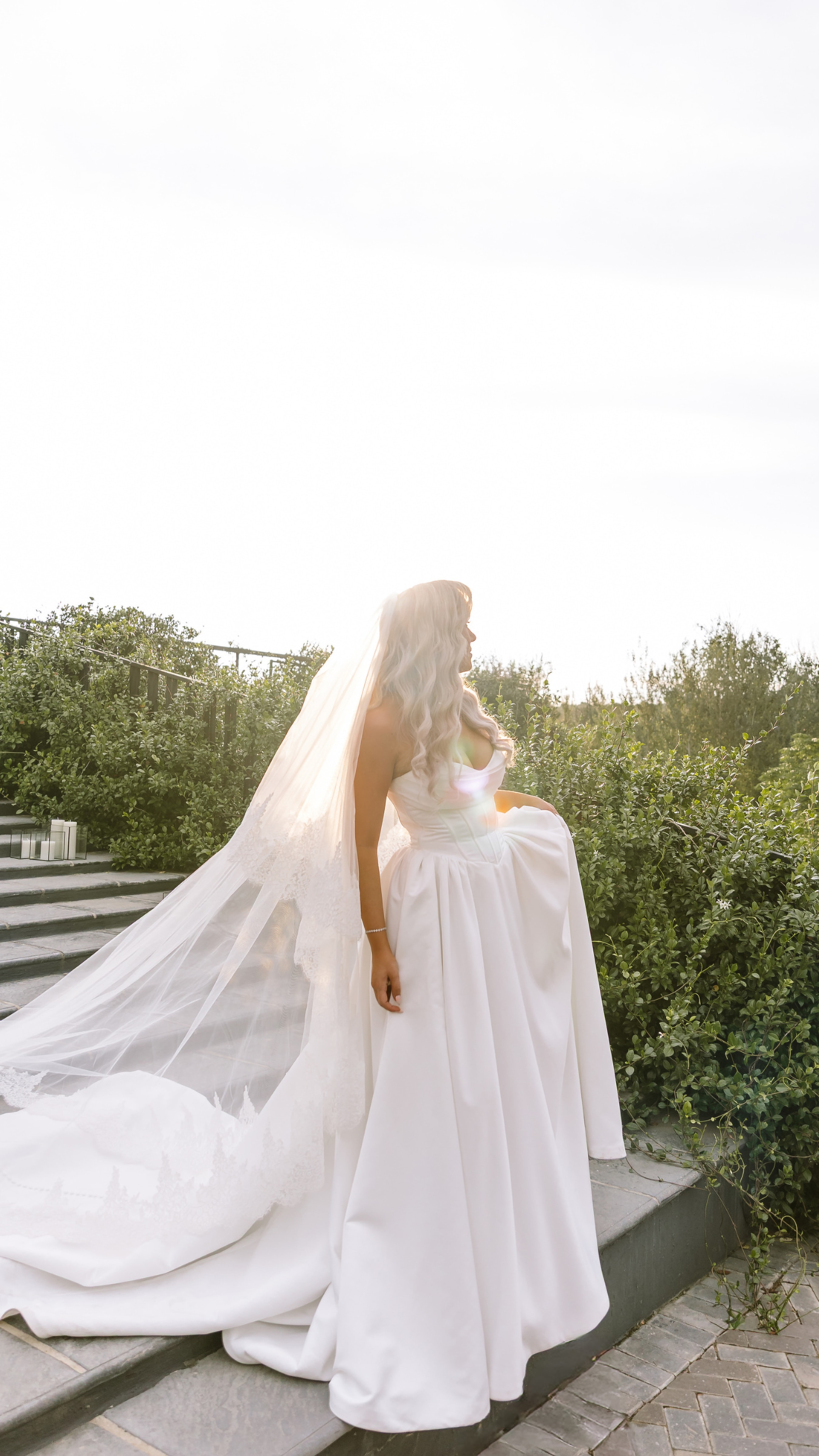 A bride in a white gown and lace veil stands on stone steps outdoors