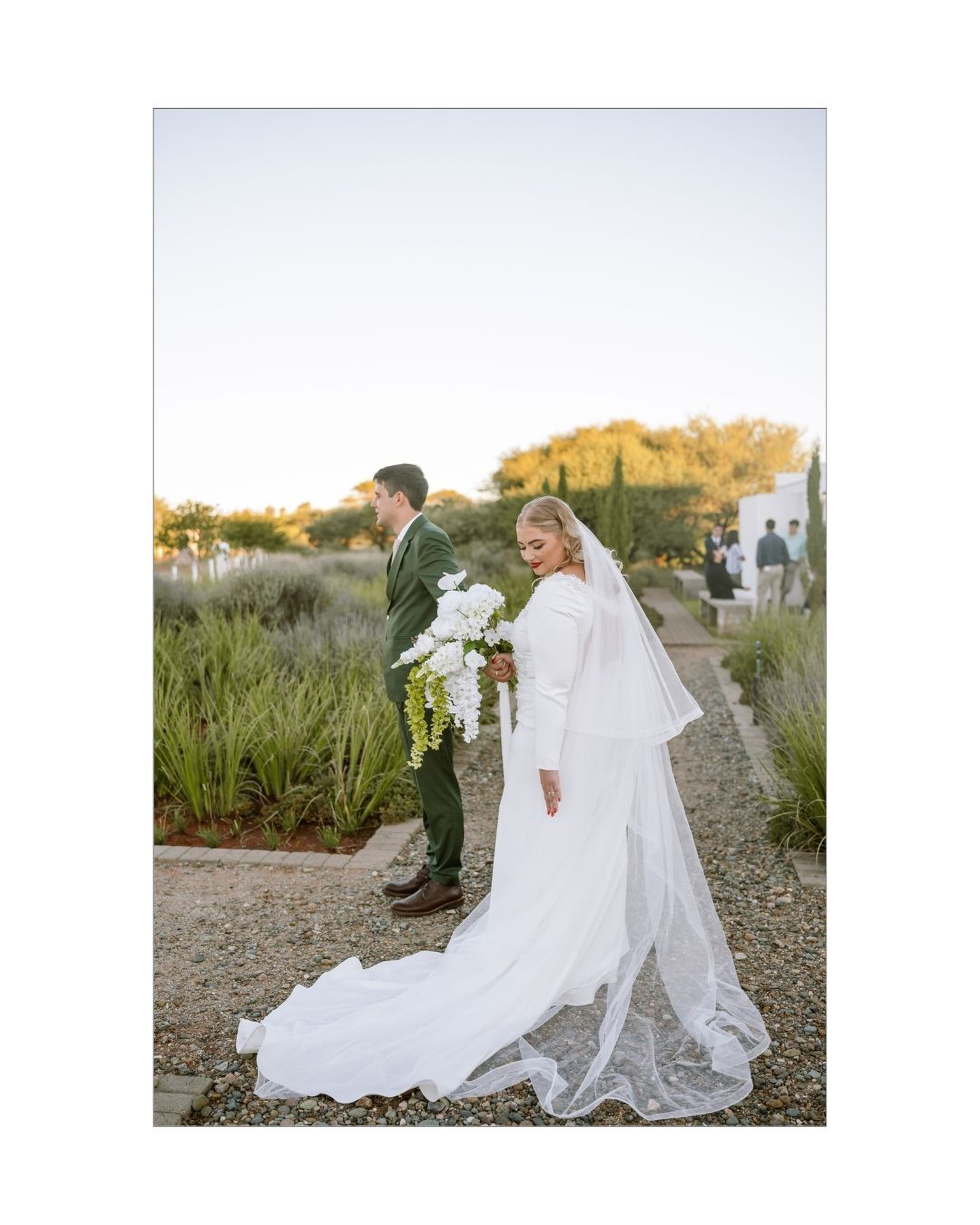 A bride in a white gown and veil holds a cascading floral bouquet near her groom