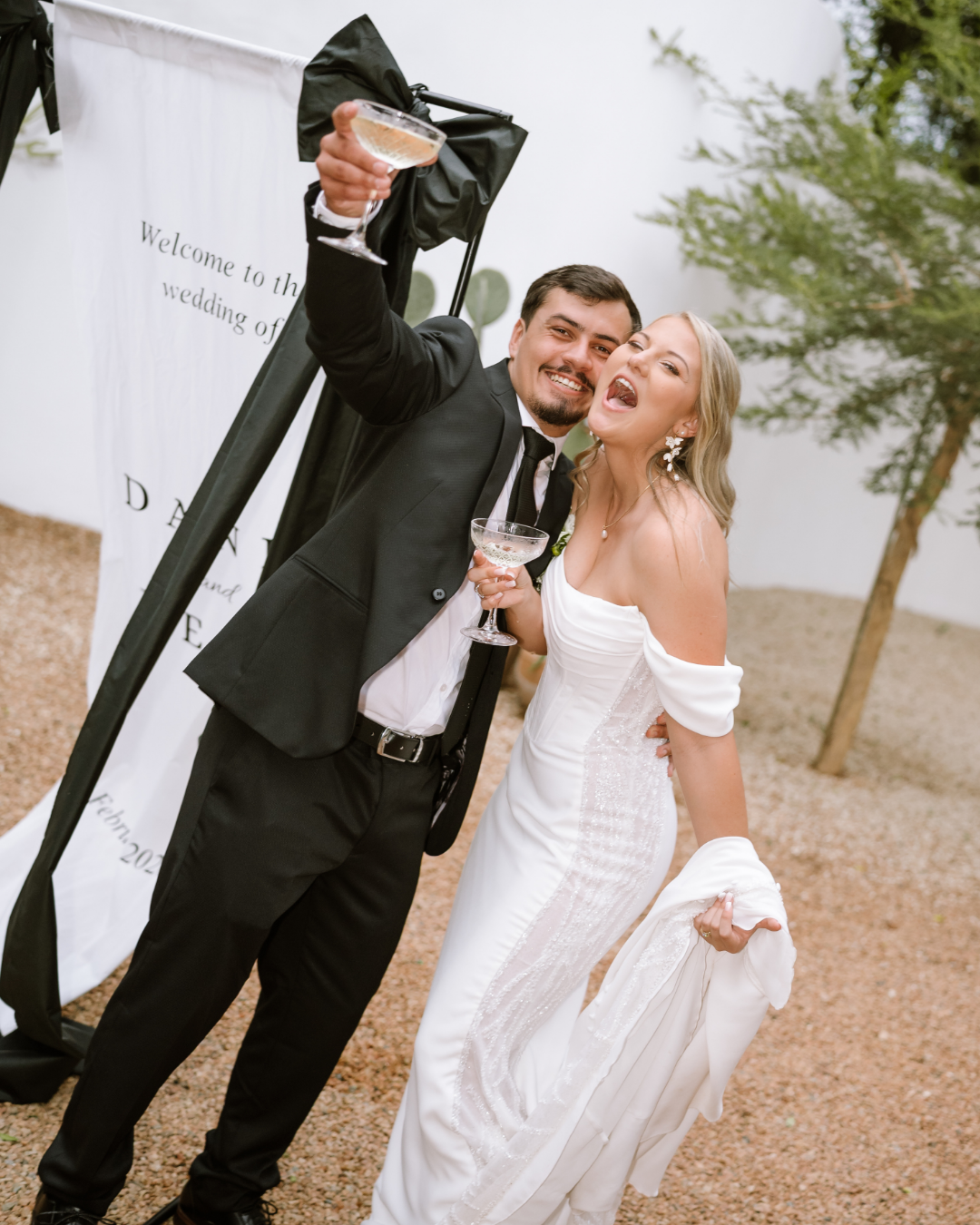 A joyful bride and groom toast with champagne glasses in front of their wedding sign