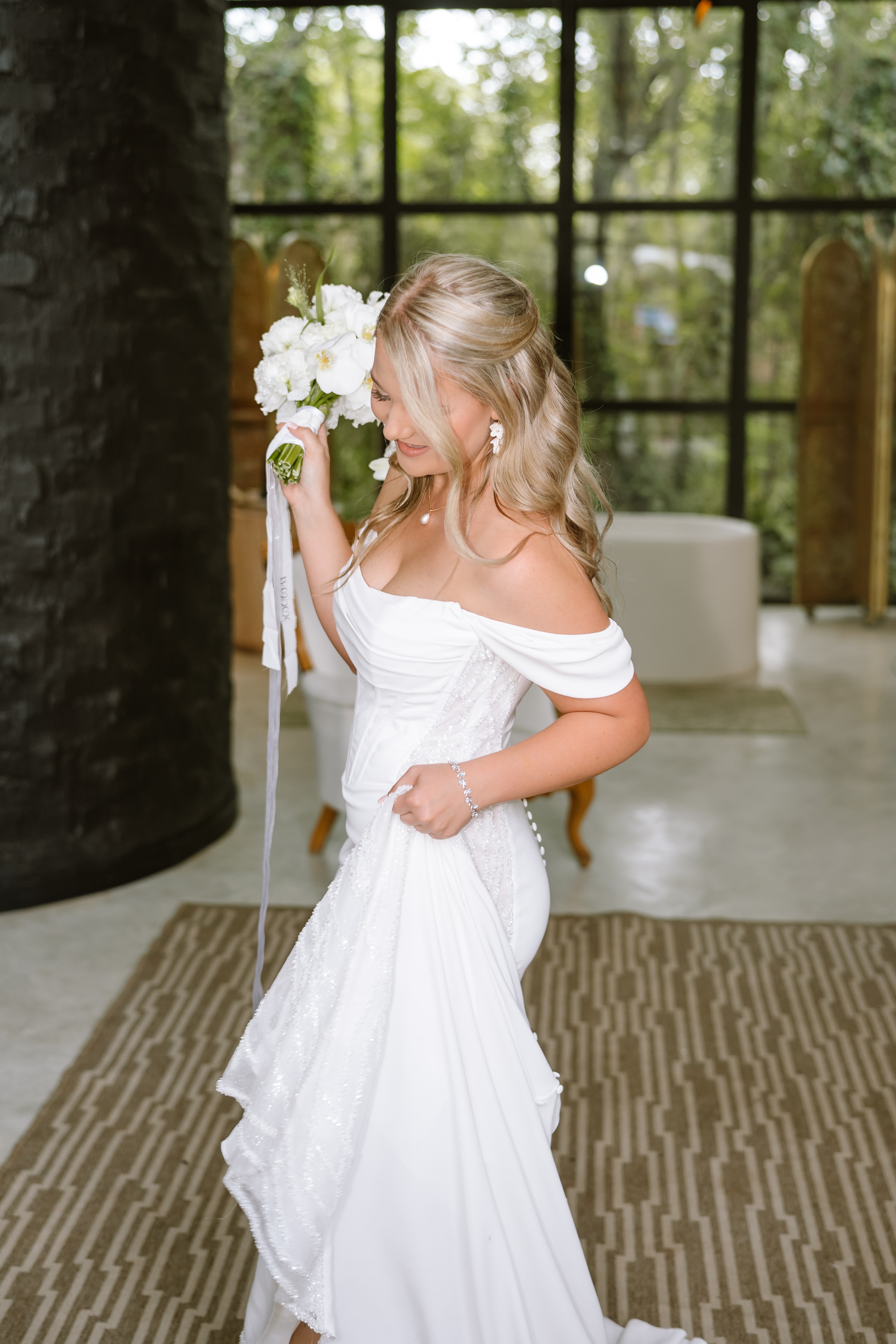 A blonde bride in a white off-the-shoulder wedding dress holds a bouquet of white flowers