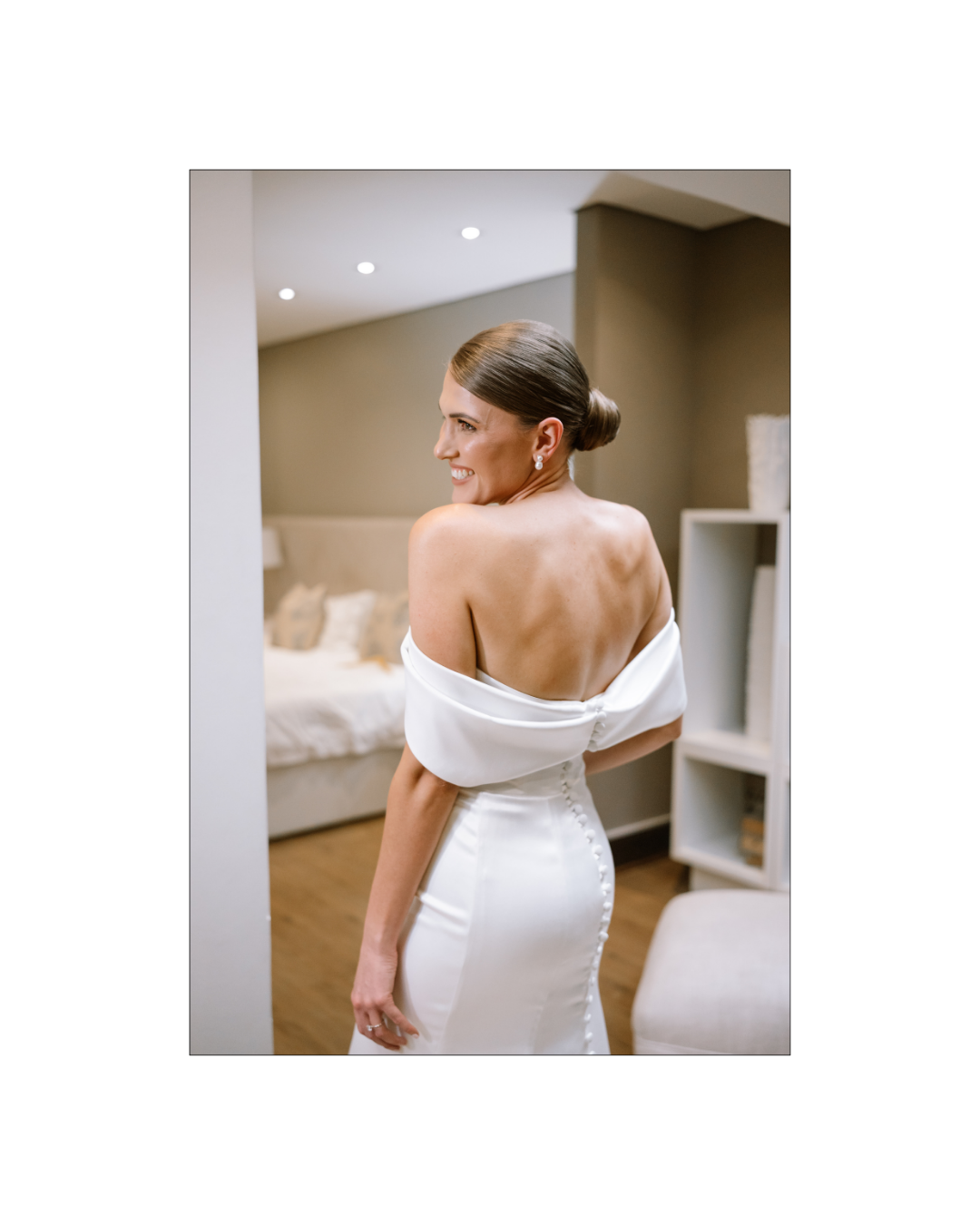 A bride in a white off-the-shoulder gown looks over her shoulder in a bedroom