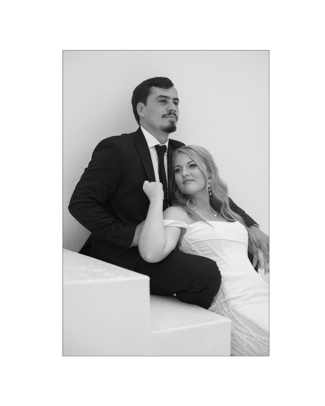 A bride in a white gown rests against her groom sitting on white stairs
