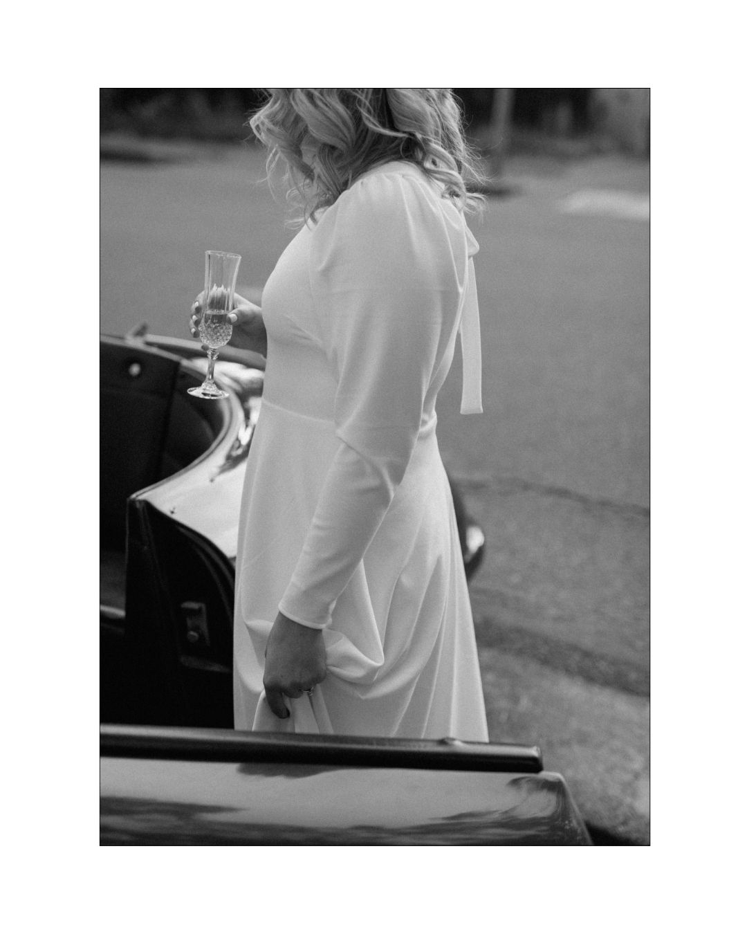 A bride in a long-sleeved white gown holding a glass of champagne near a car