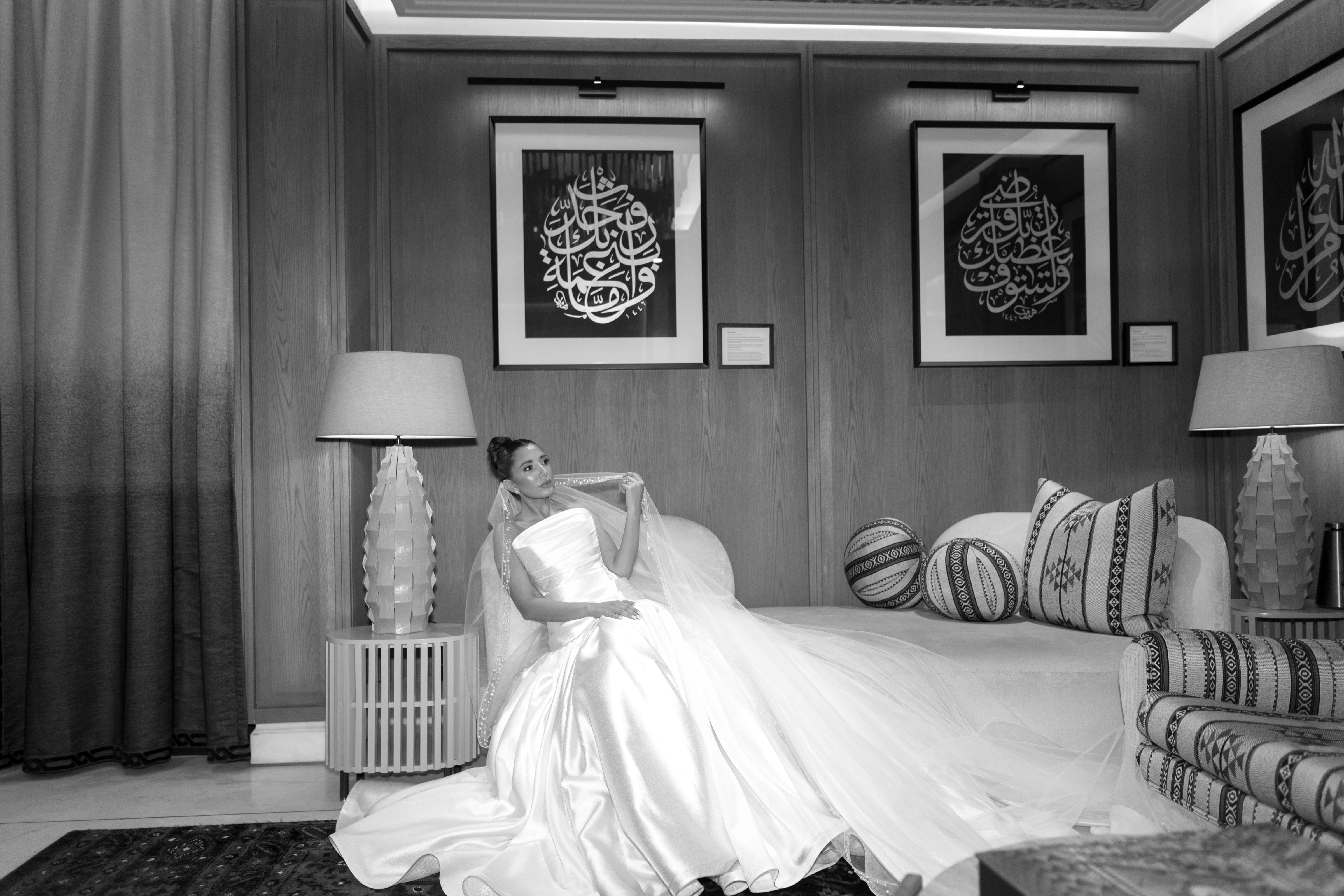 A bride in a white wedding gown sits elegantly on a sofa under framed calligraphy