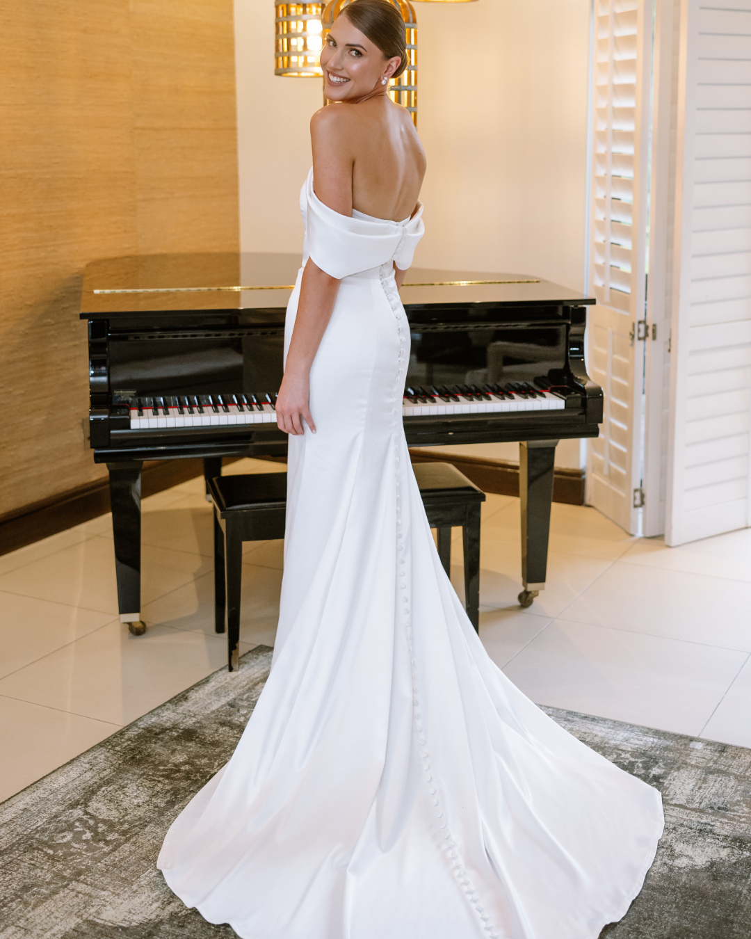 A smiling bride wearing a white off-the-shoulder wedding dress stands before a grand piano