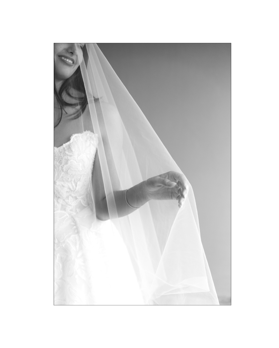 A smiling bride wearing a delicate lace wedding dress holds her elegant sheer veil gracefully