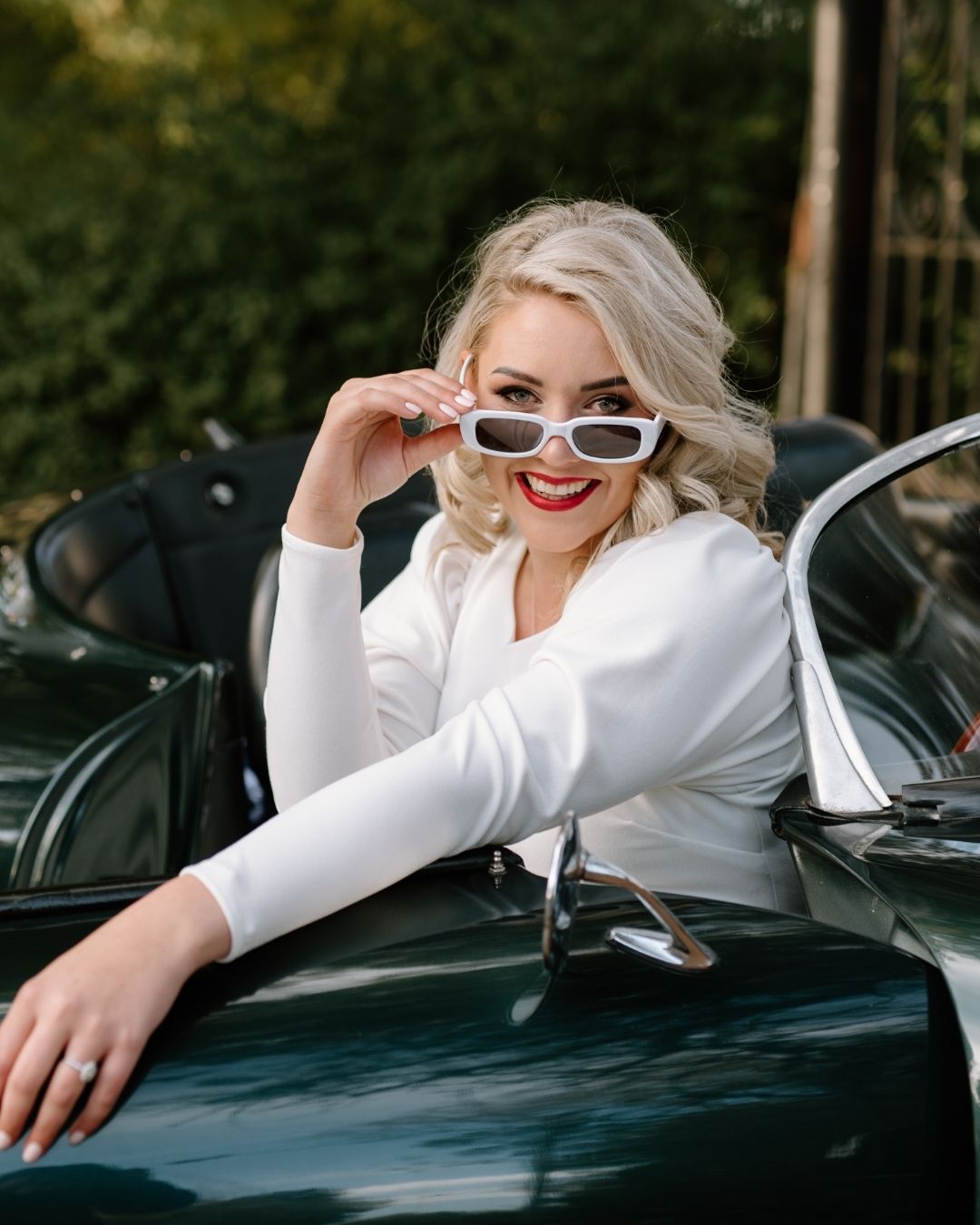 A smiling blonde bride wearing white sunglasses while sitting in a vintage green convertible car