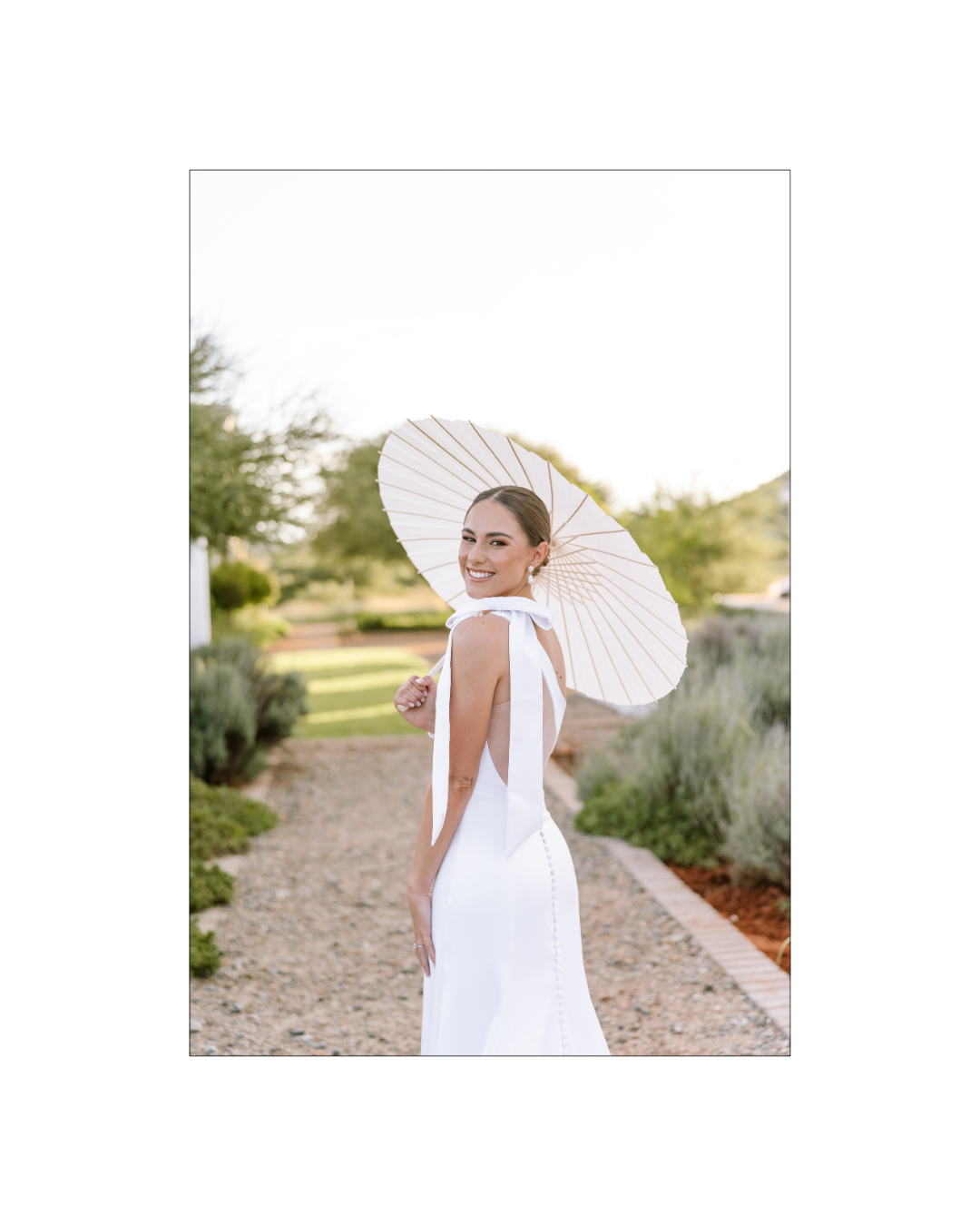 A smiling bride in a white gown holds a parasol on a gravel path