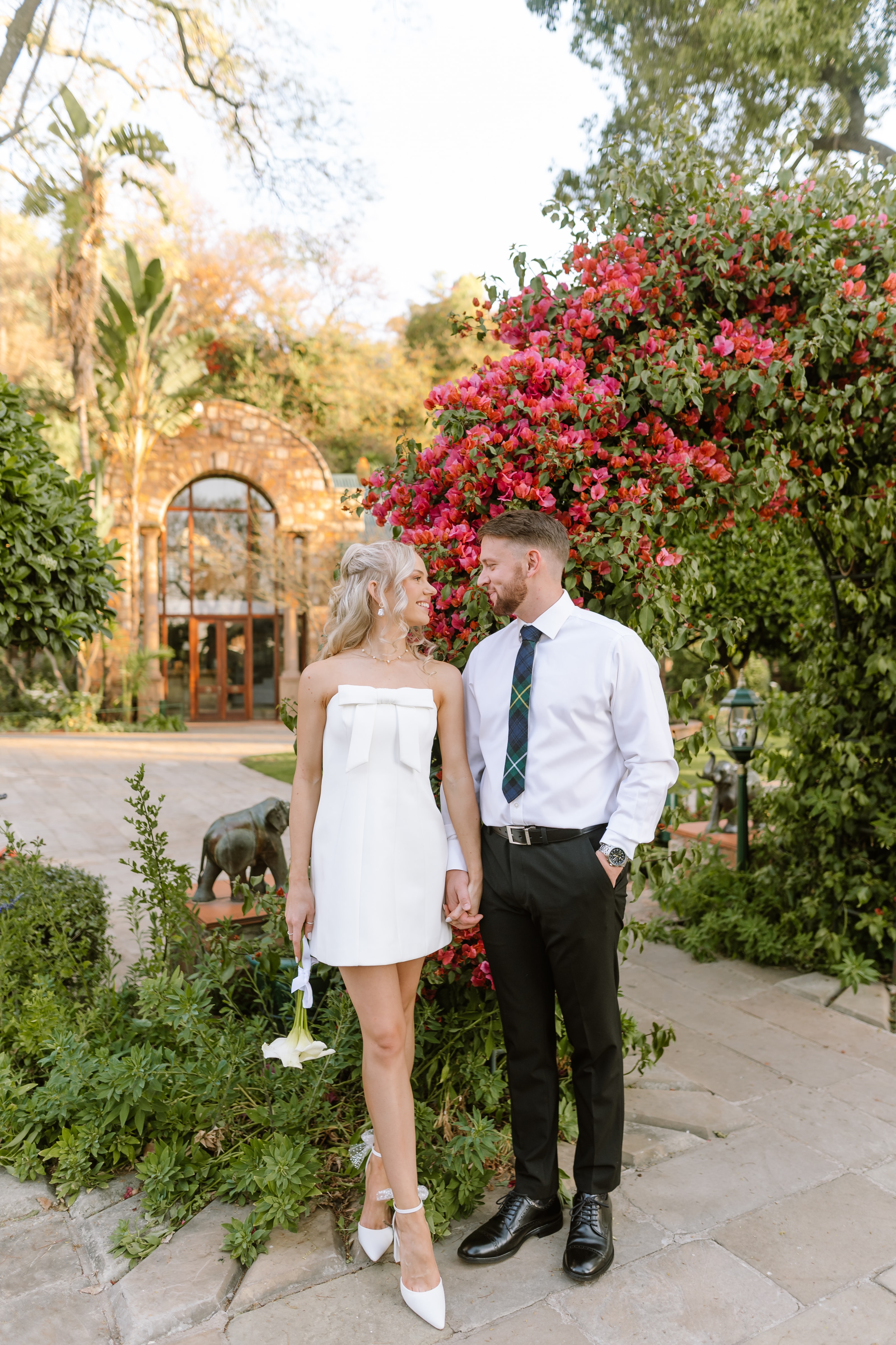 A stylish couple stands together in a lush garden near a stone archway during their wedding