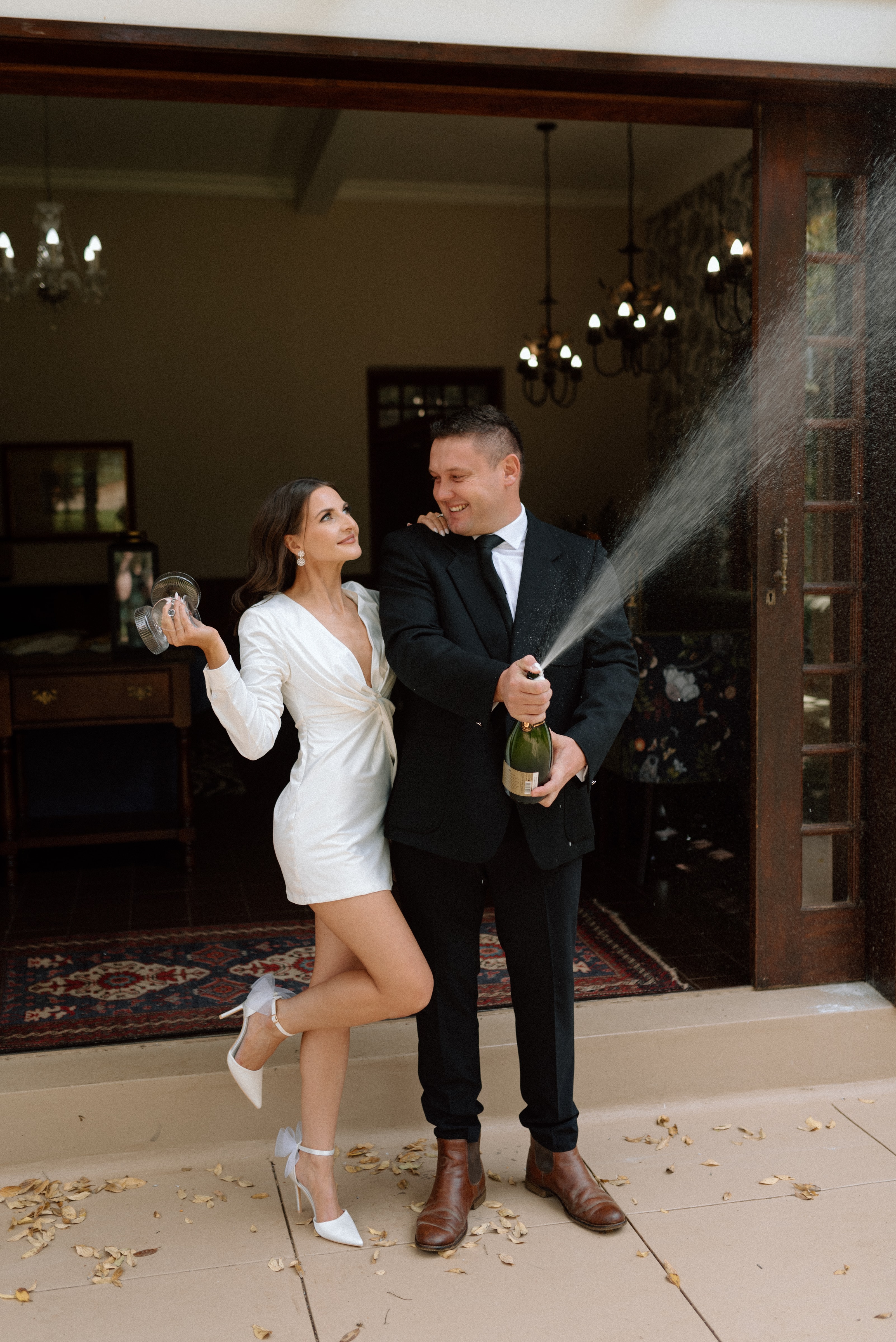 A stylish bride and groom celebrating with a champagne pop outside a luxury estate venue