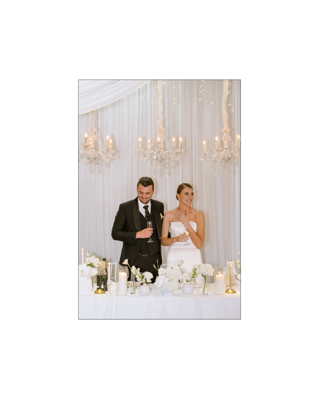 A bride and groom stand behind a decorated wedding table holding champagne flutes together