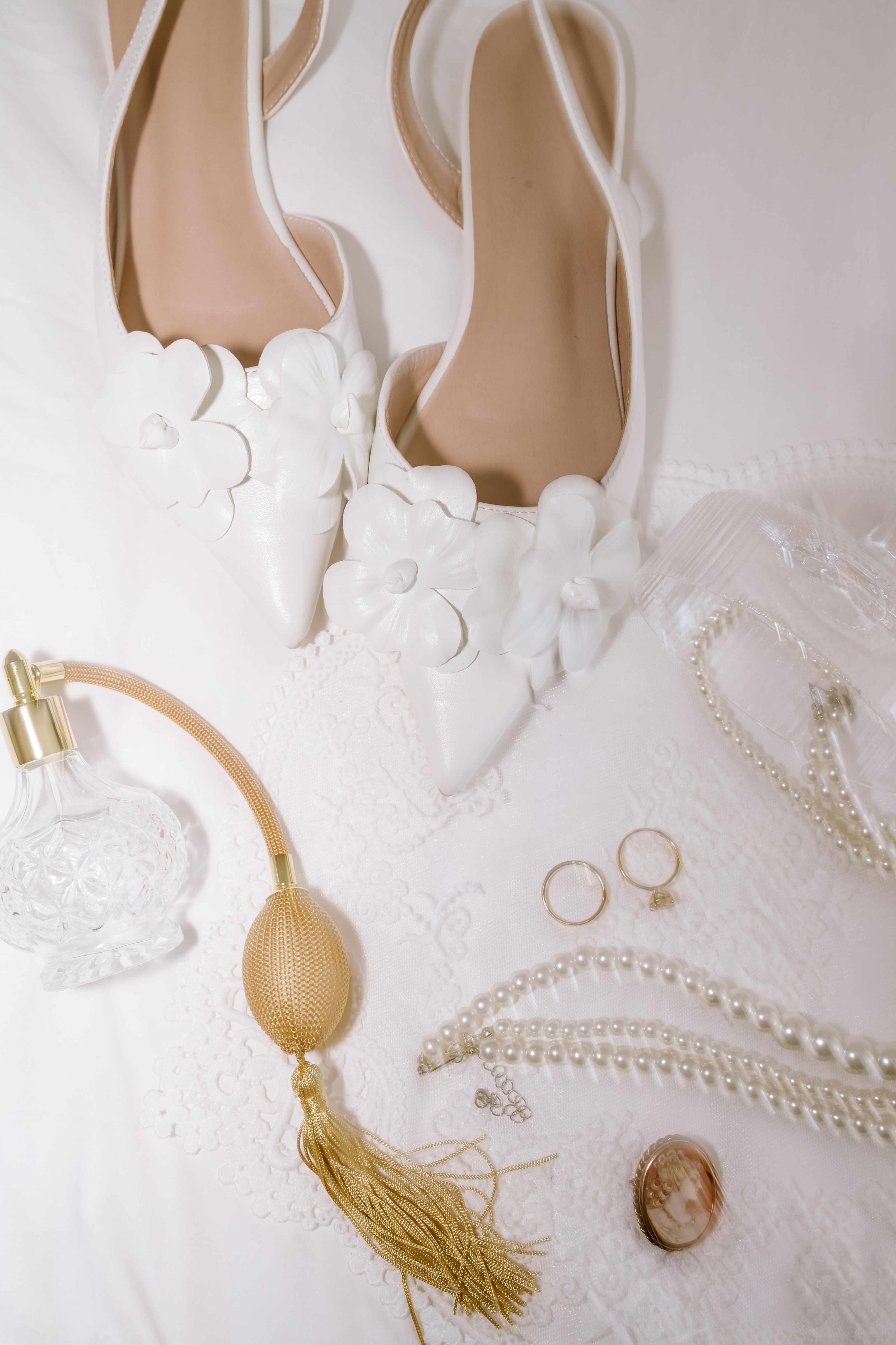 Elegant white wedding shoes arranged alongside pearl necklaces, gold rings, and a vintage perfume bottle