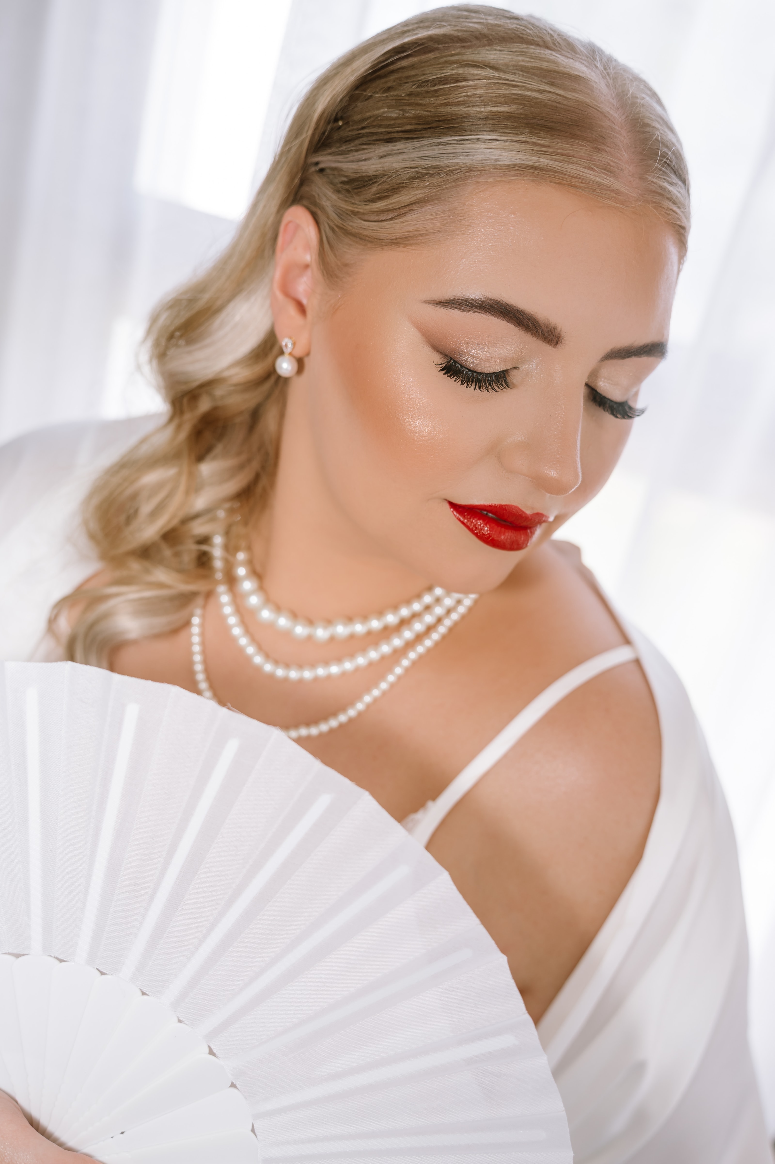 A glamorous bride with blonde hair holding a white fan and wearing pearls