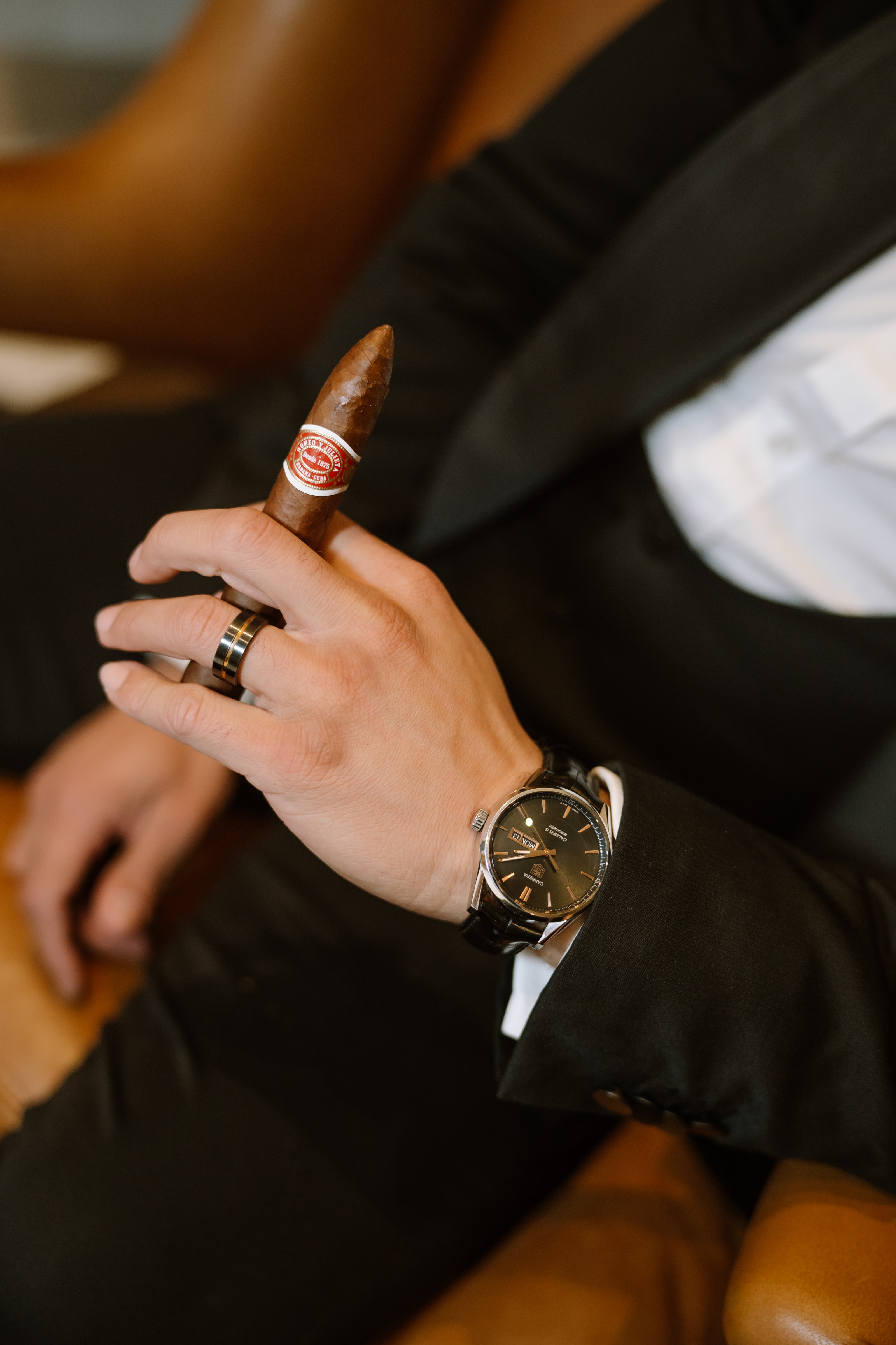A groom in a black suit holds a cigar while wearing a luxury wristwatch