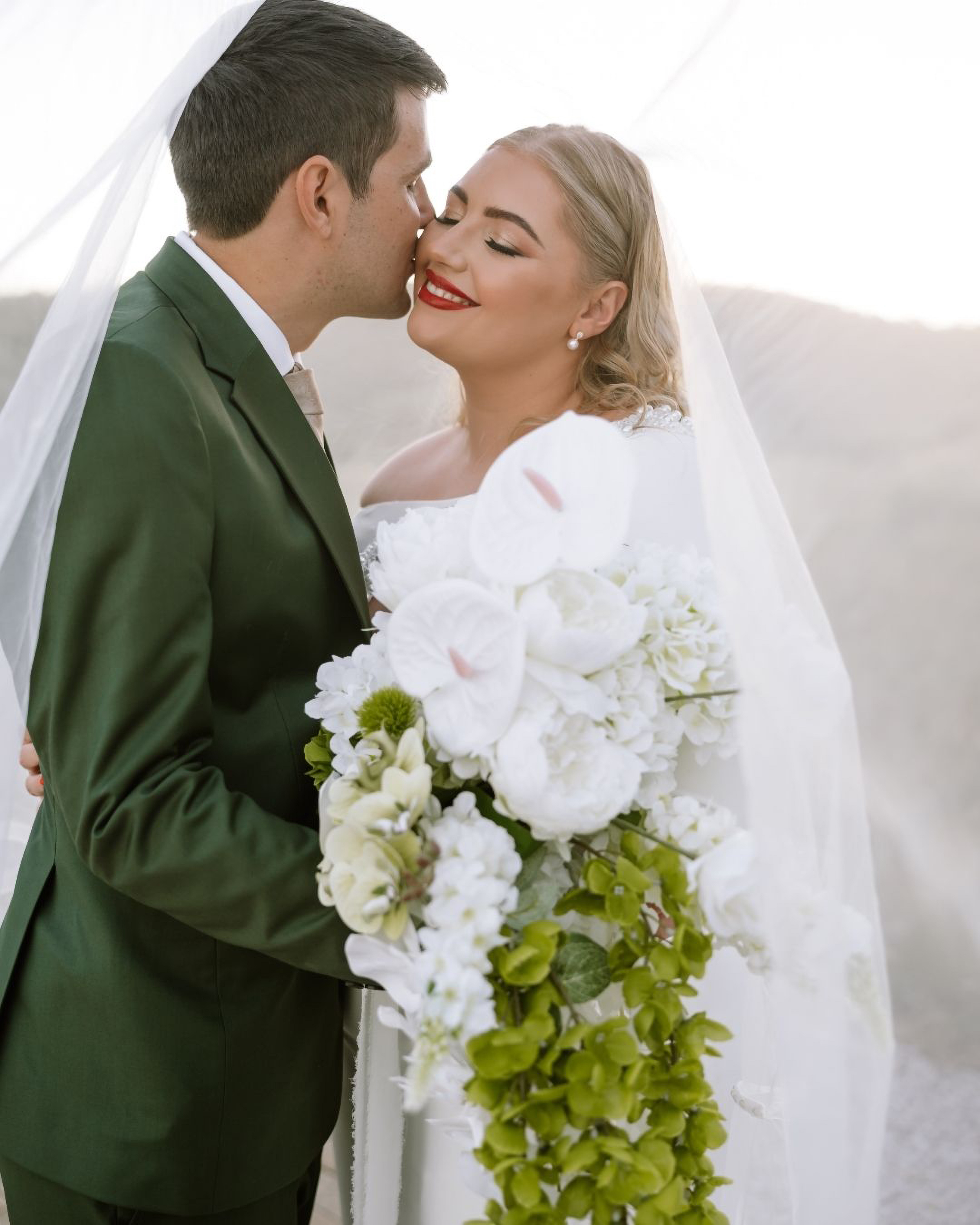 Groom in a green suit kisses smiling bride under her veil holding white flowers