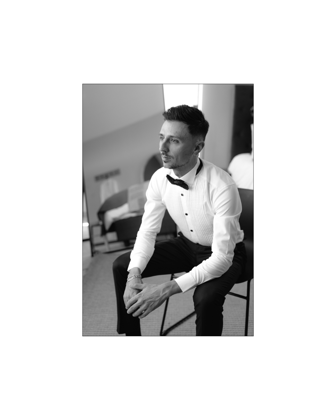 A stylish groom in a crisp white tuxedo shirt and bow tie sits pensively indoors