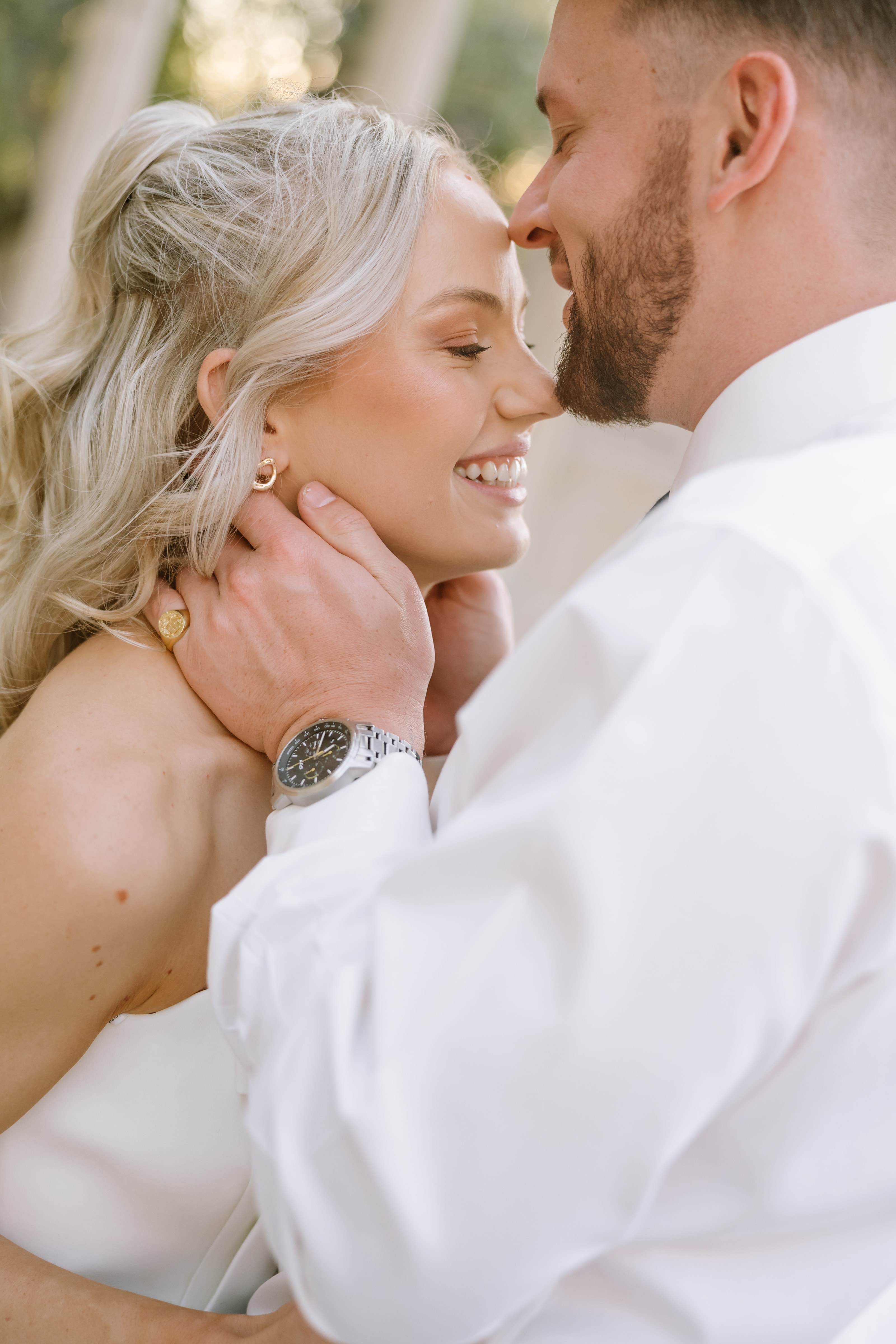 A groom gently cradles his smiling bride's face during an intimate outdoor wedding portrait session