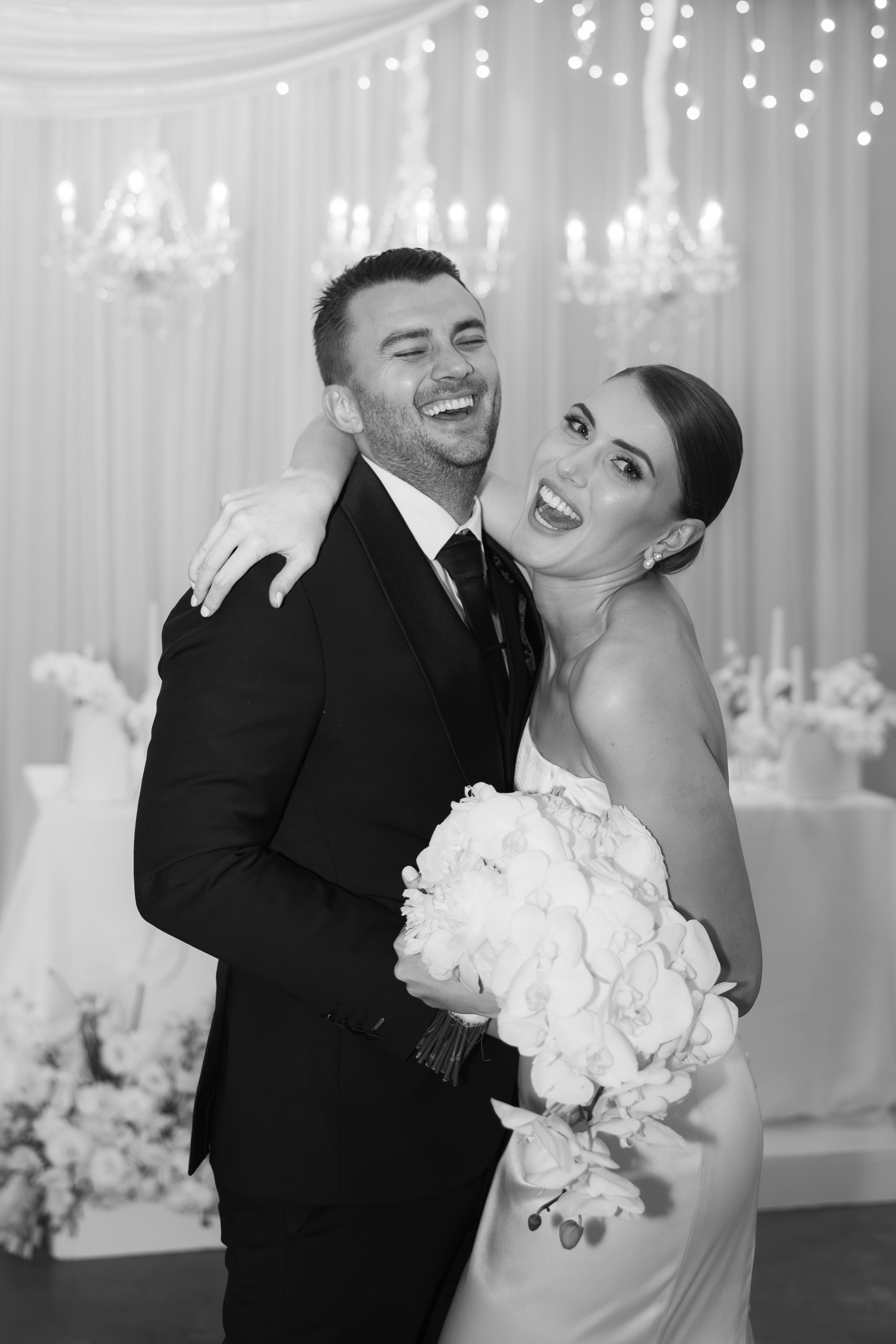 A joyful bride and groom laughing together while holding a large white floral bouquet