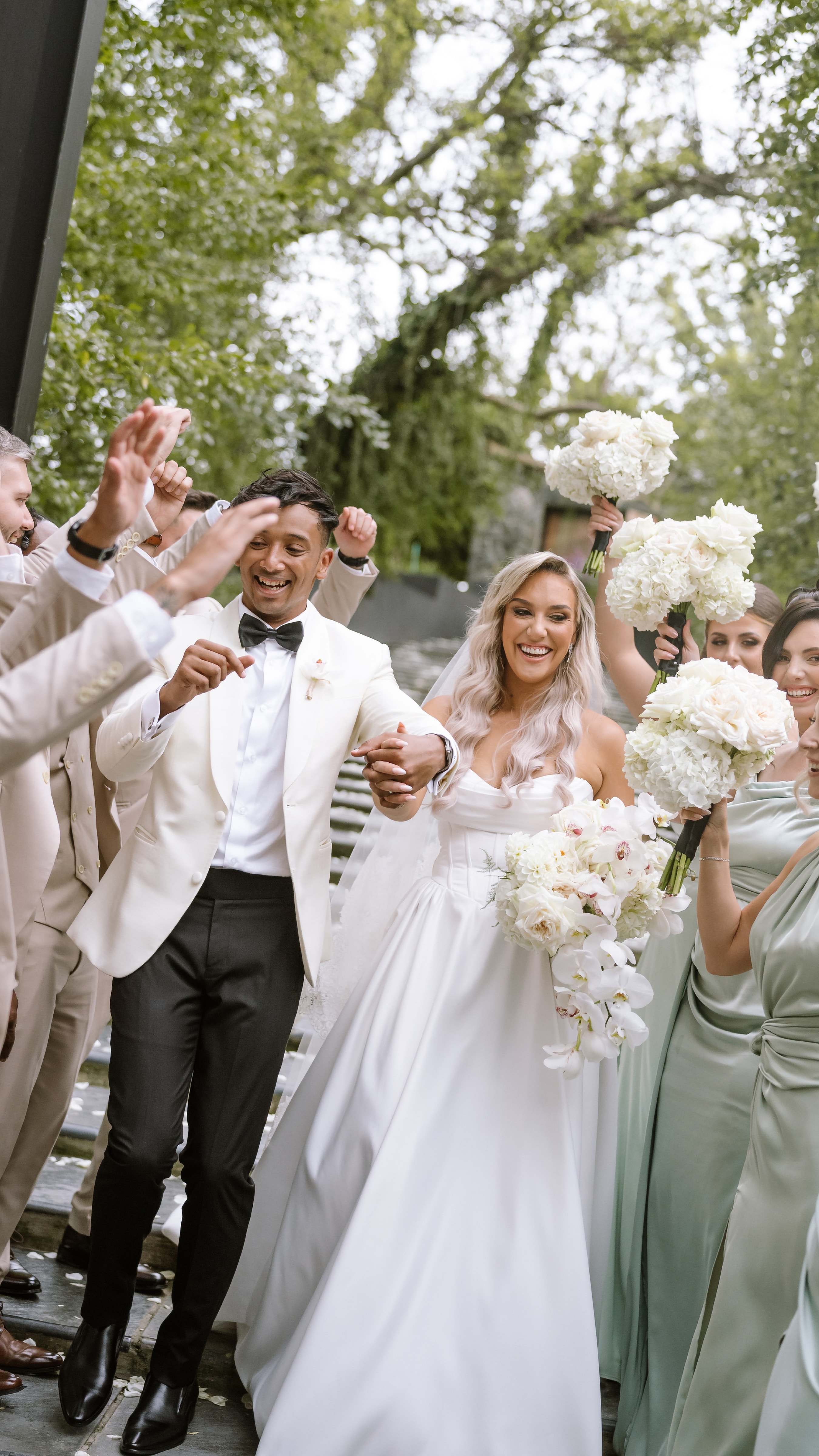 A joyful bride and groom walk hand in hand surrounded by their happy wedding party