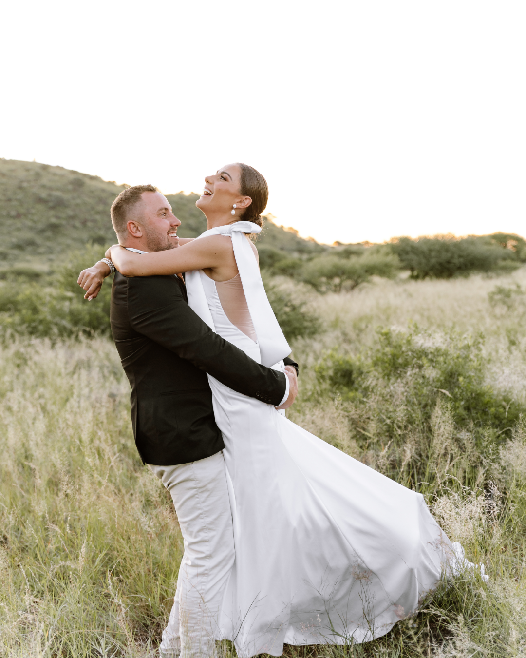 A joyful bride in a white wedding gown laughing as her groom lifts her up outdoors