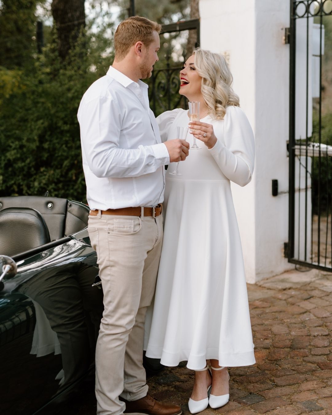 A joyful couple in white wedding attire shares a celebratory drink beside a vintage car