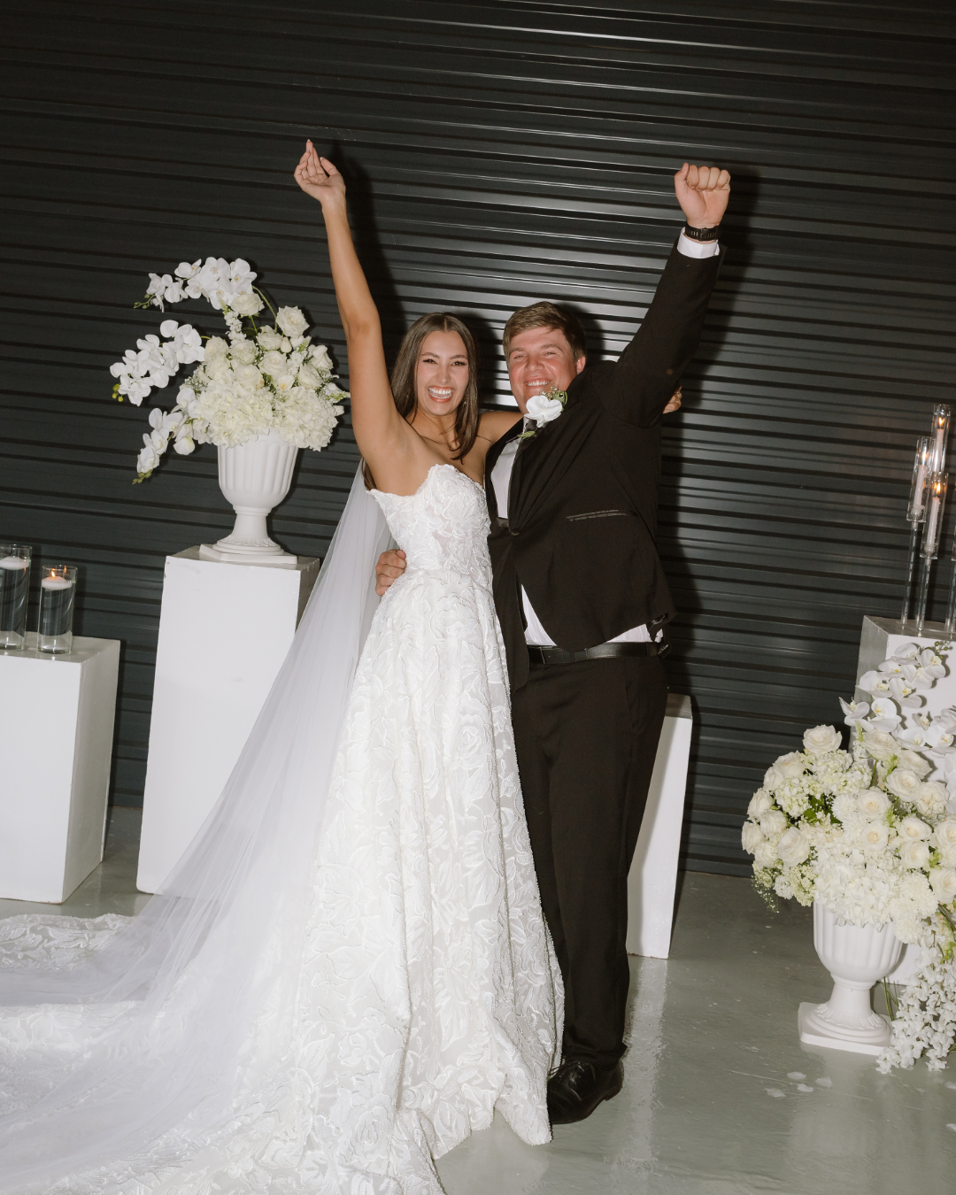 A joyful bride and groom celebrate their marriage with arms raised against a modern backdrop