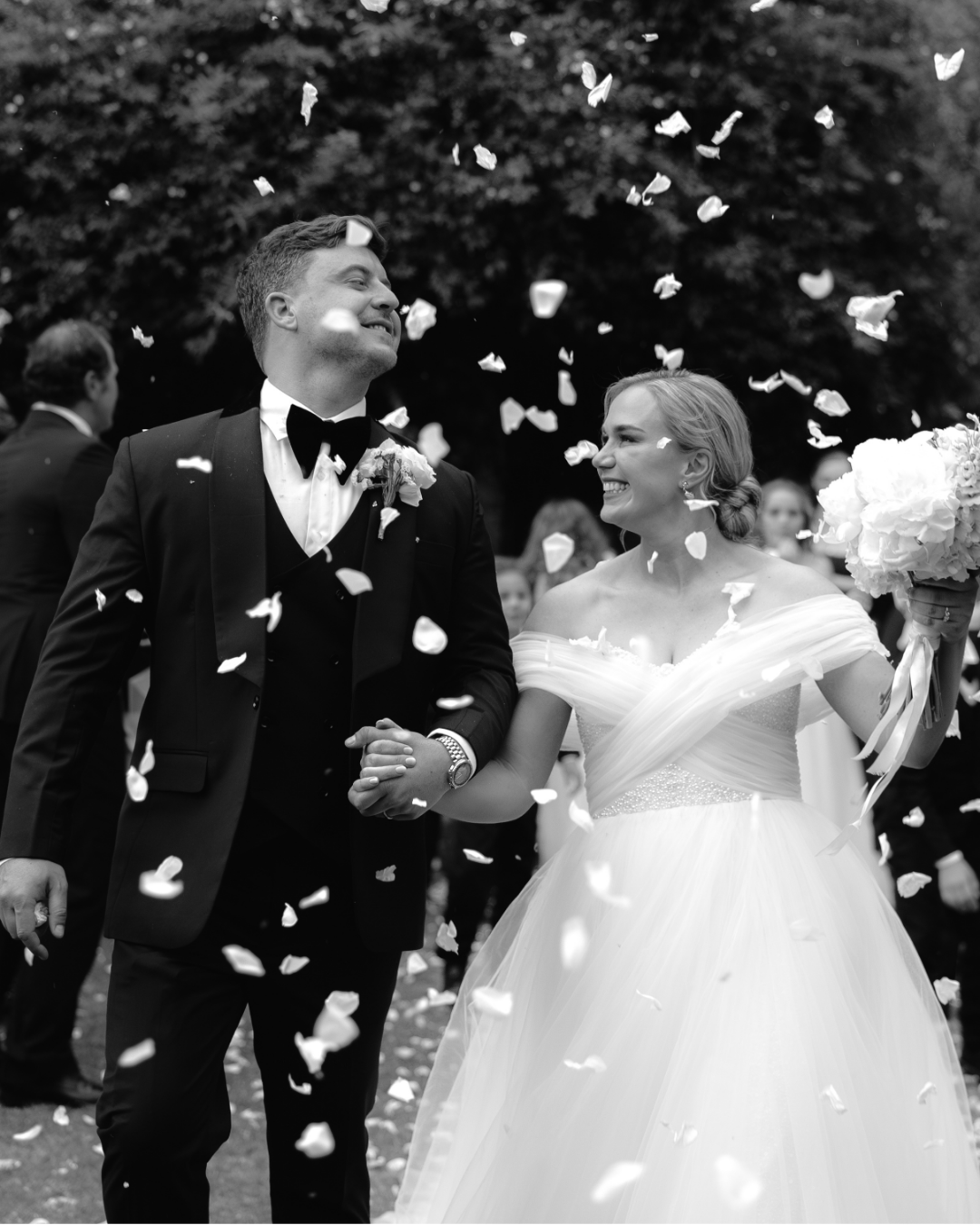 A joyful bride and groom walk together as guests shower them with white flower petals
