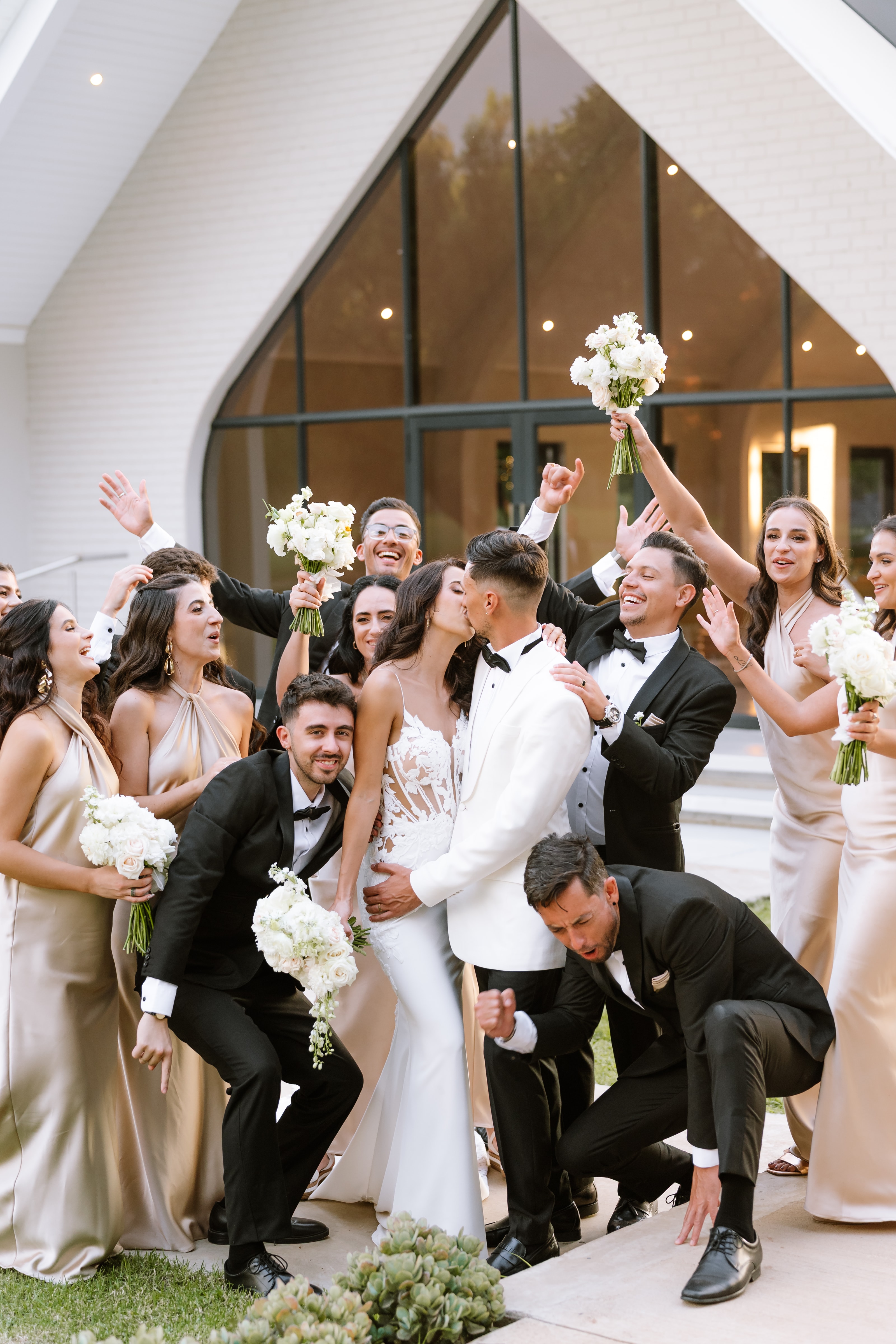 A joyful wedding party celebrates with the bride and groom kissing in front of a modern venue