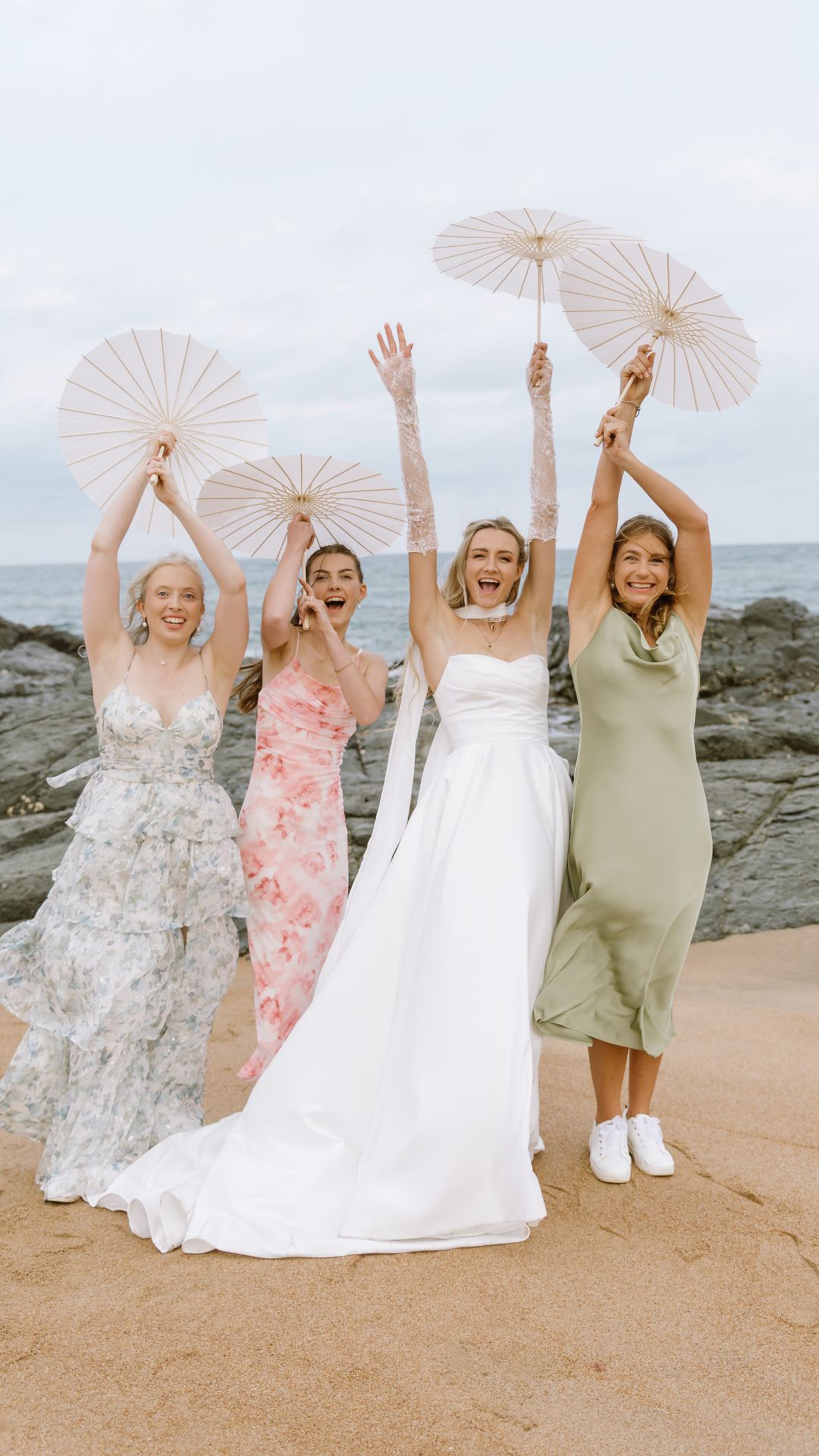 A laughing bride and three bridesmaids hold white parasols on a sandy beach