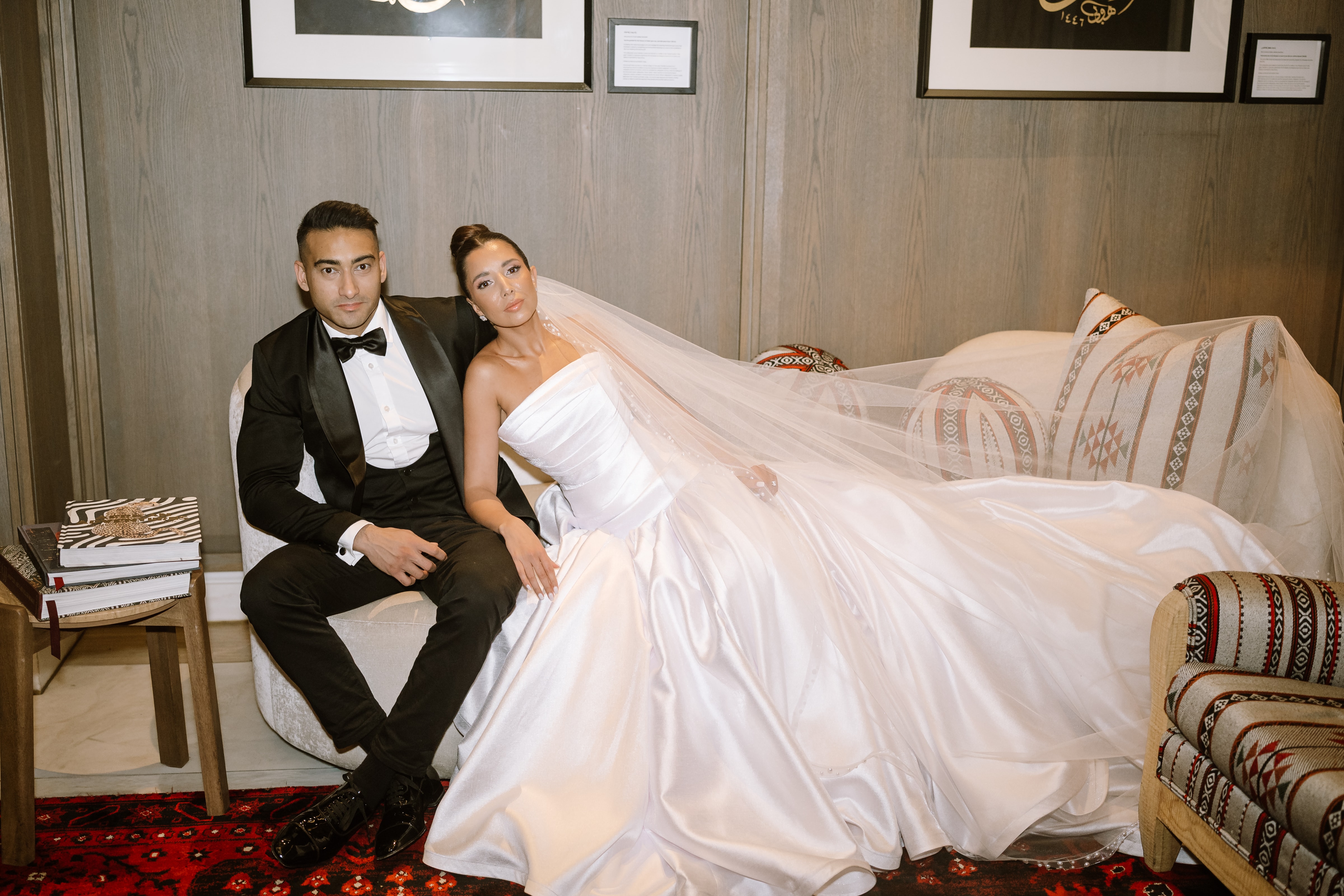 A newlywed couple poses elegantly on a cream sofa inside a sophisticated, warmly lit room