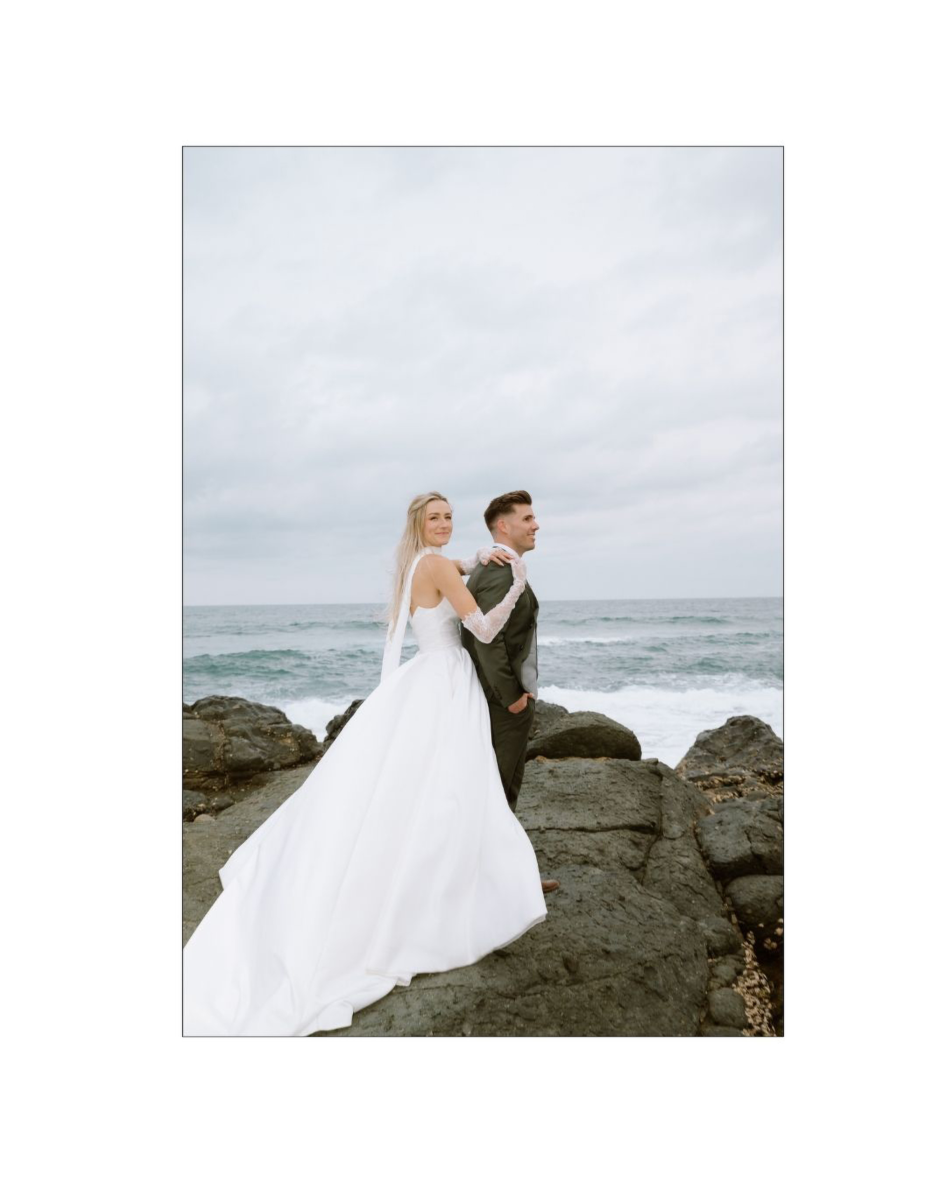 A newlywed couple poses on rocky shores overlooking a moody ocean under an overcast sky