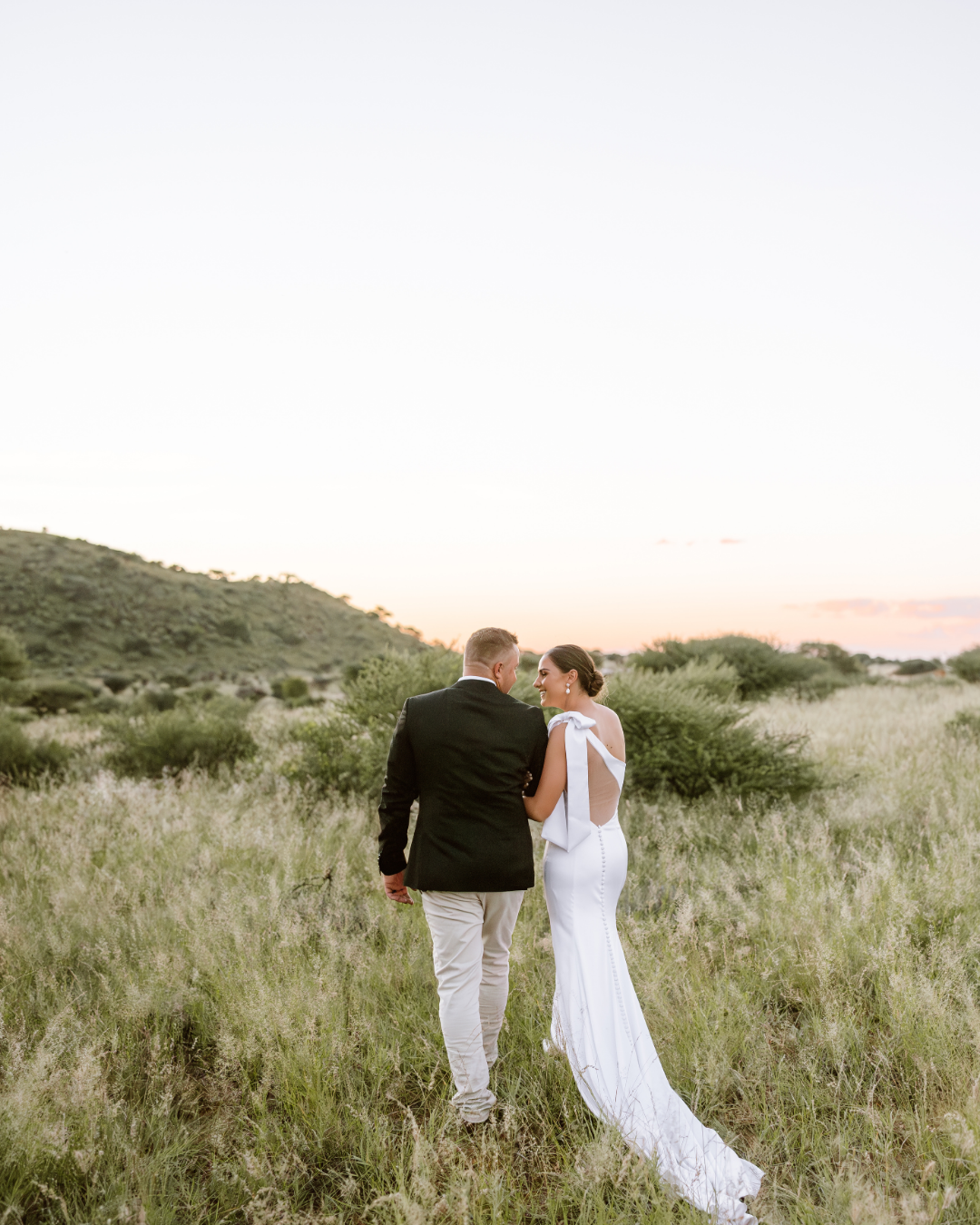 A newlywed couple walks through tall golden grass in a scenic South African landscape