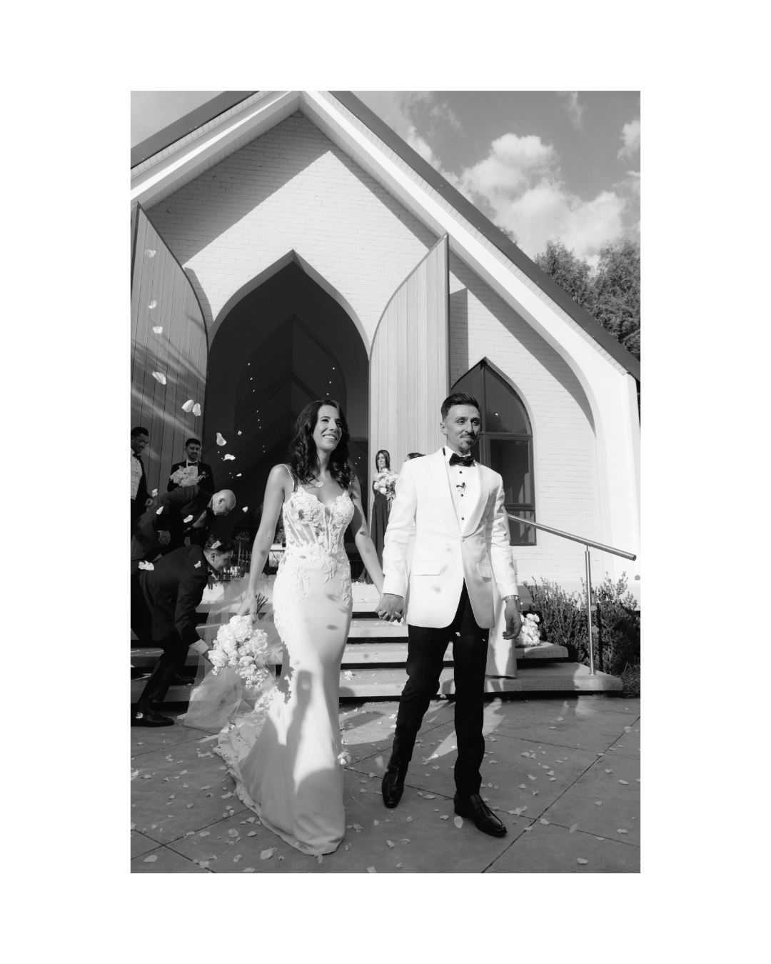 A newlywed couple walks hand in hand from a modern chapel as guests throw white petals