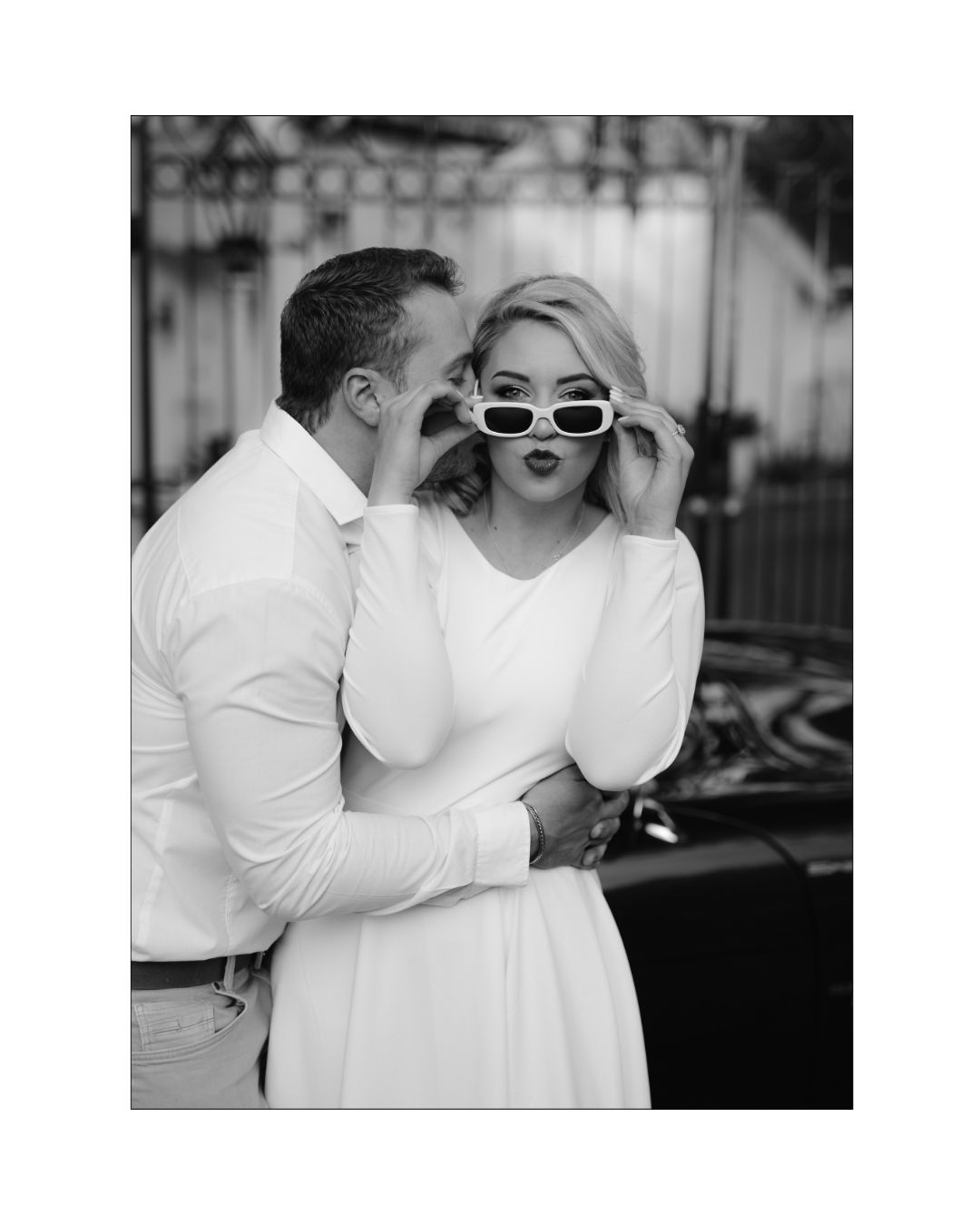 A couple in white attire embraces as the woman playfully adjusts her white sunglasses