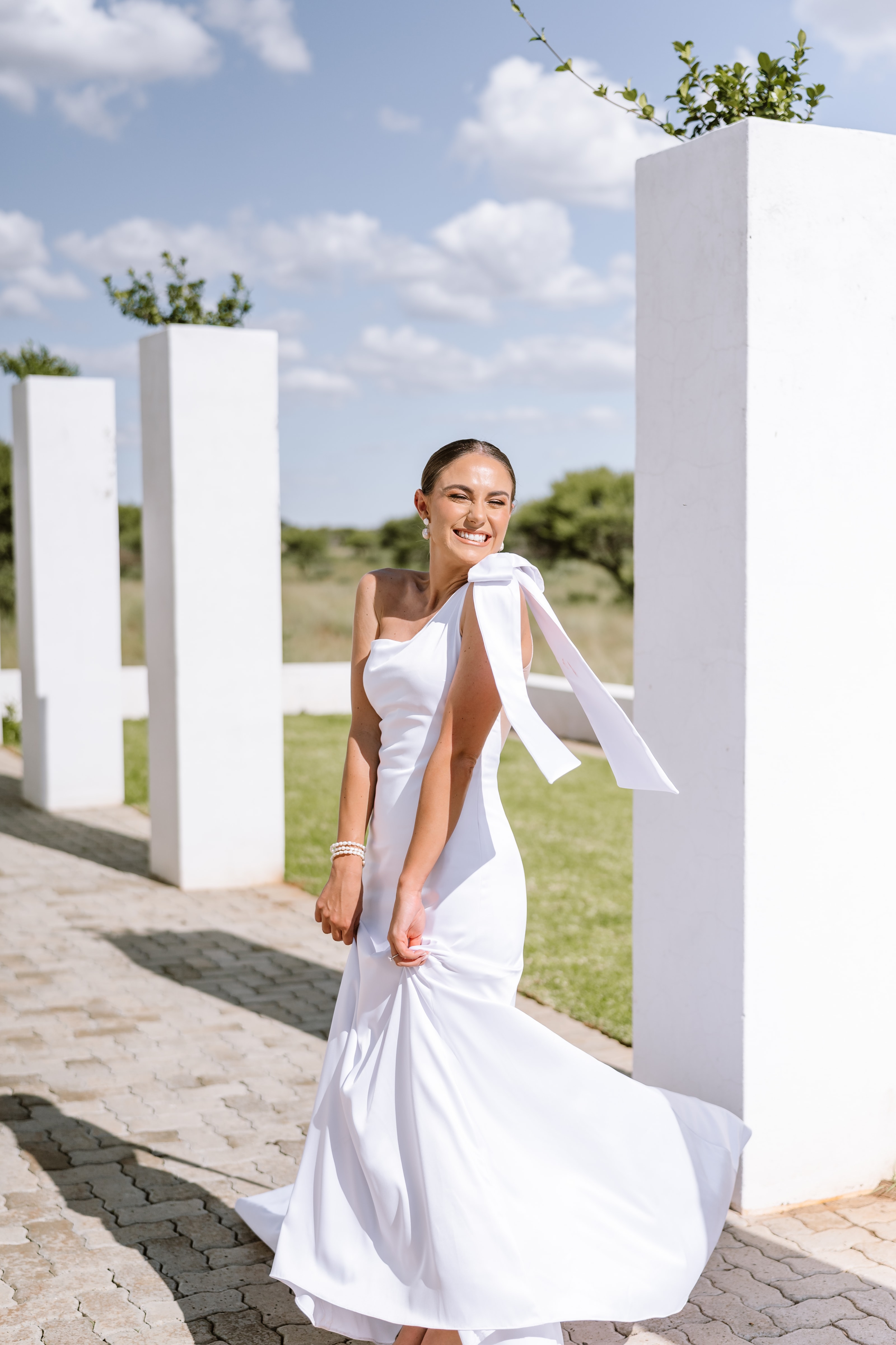 A joyful bride in a white one-shoulder gown posing between modern stone pillars outdoors